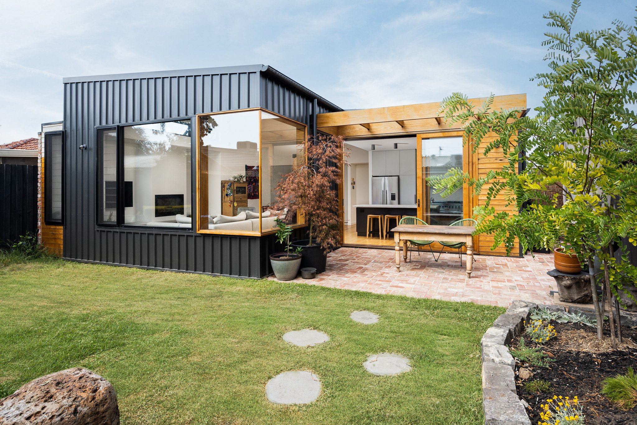 Modern house with black metal siding, large windows, and a wooden deck with outdoor furniture, surrounded by a well-maintained lawn and garden.