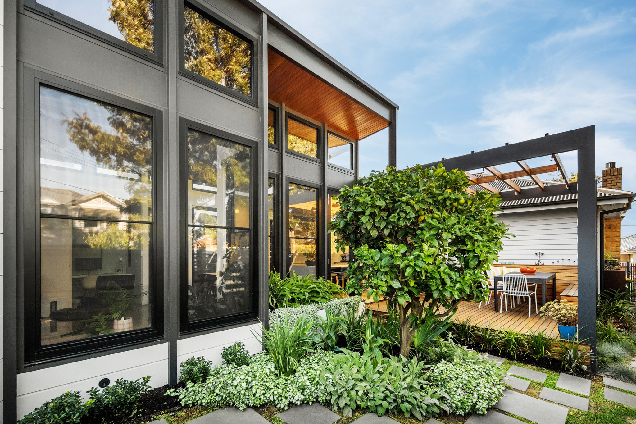 Modern house with black framed windows and a garden with various green plants and a small tree, a wooden deck with outdoor furniture, and a pergola structure against a blue sky.