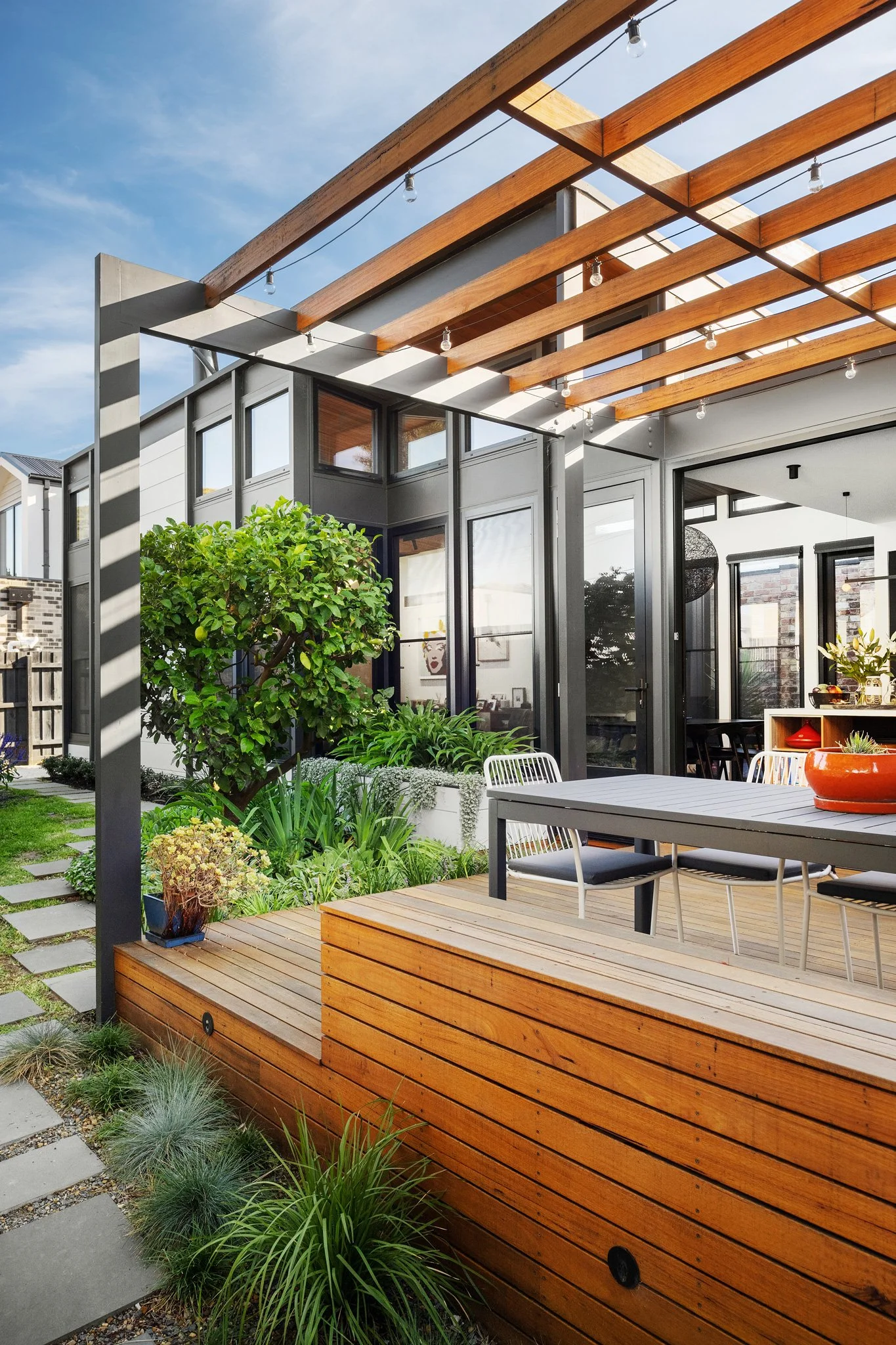 Modern backyard patio with wooden deck, outdoor dining table, black chairs, lush green plants, and a pergola with string lights