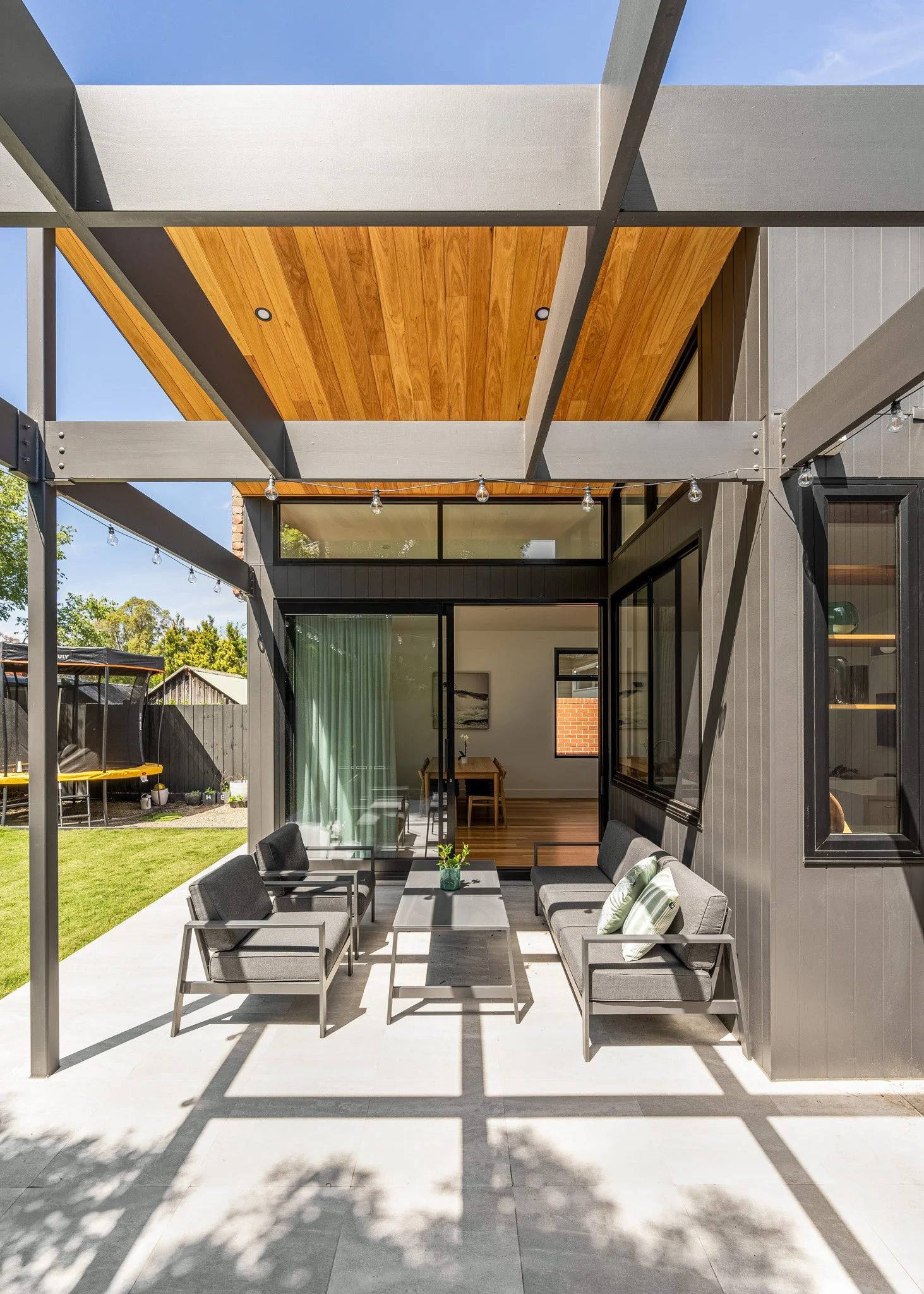 Modern outdoor patio with gray cushioned chairs and a coffee table, attached to a house with black siding and large windows, overlooking a backyard with green grass and a trampoline.