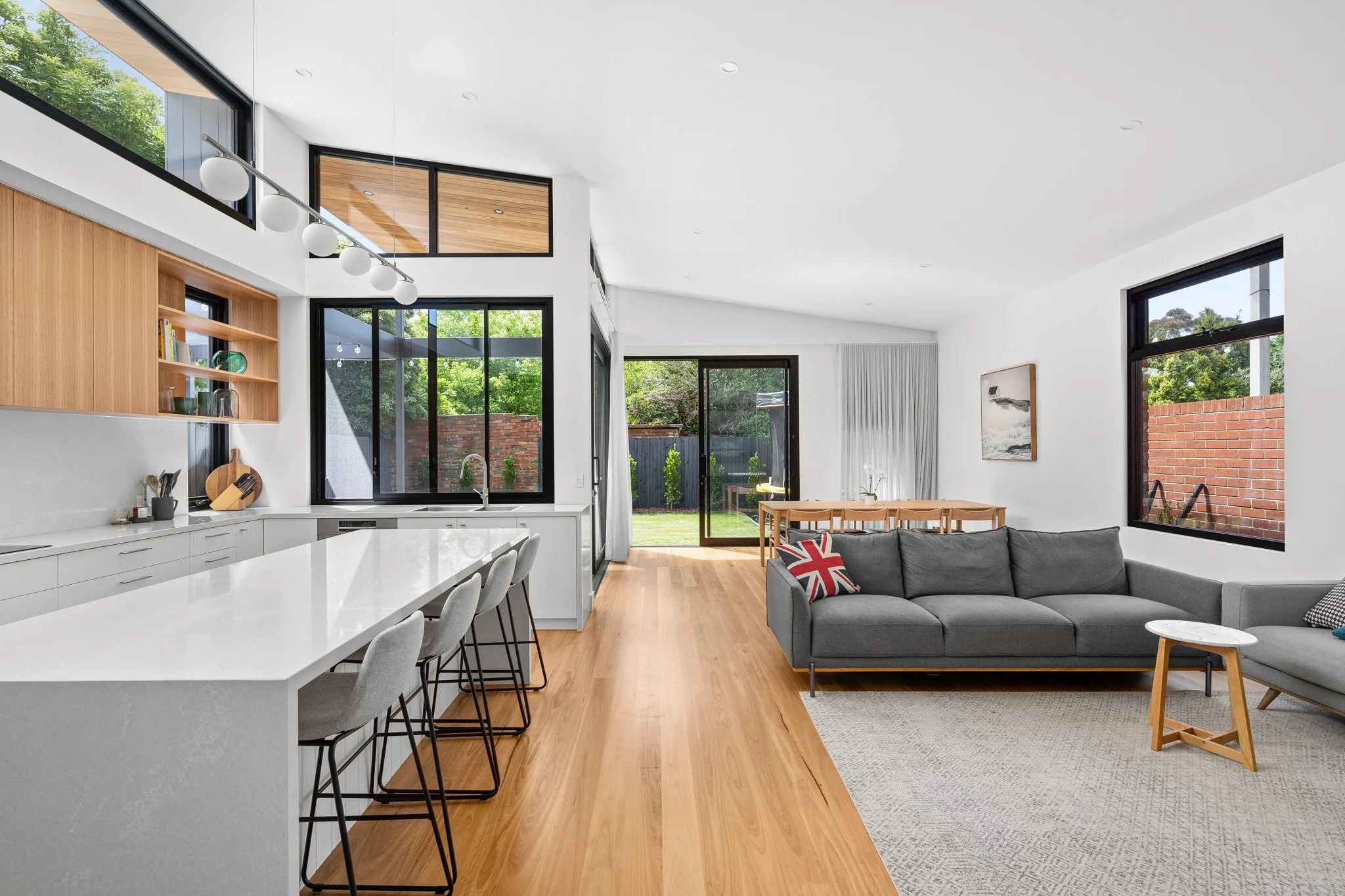Open-concept living room and kitchen with white walls, wooden flooring, large windows, a gray sofa with a Union Jack pillow, and a dining table with chairs.