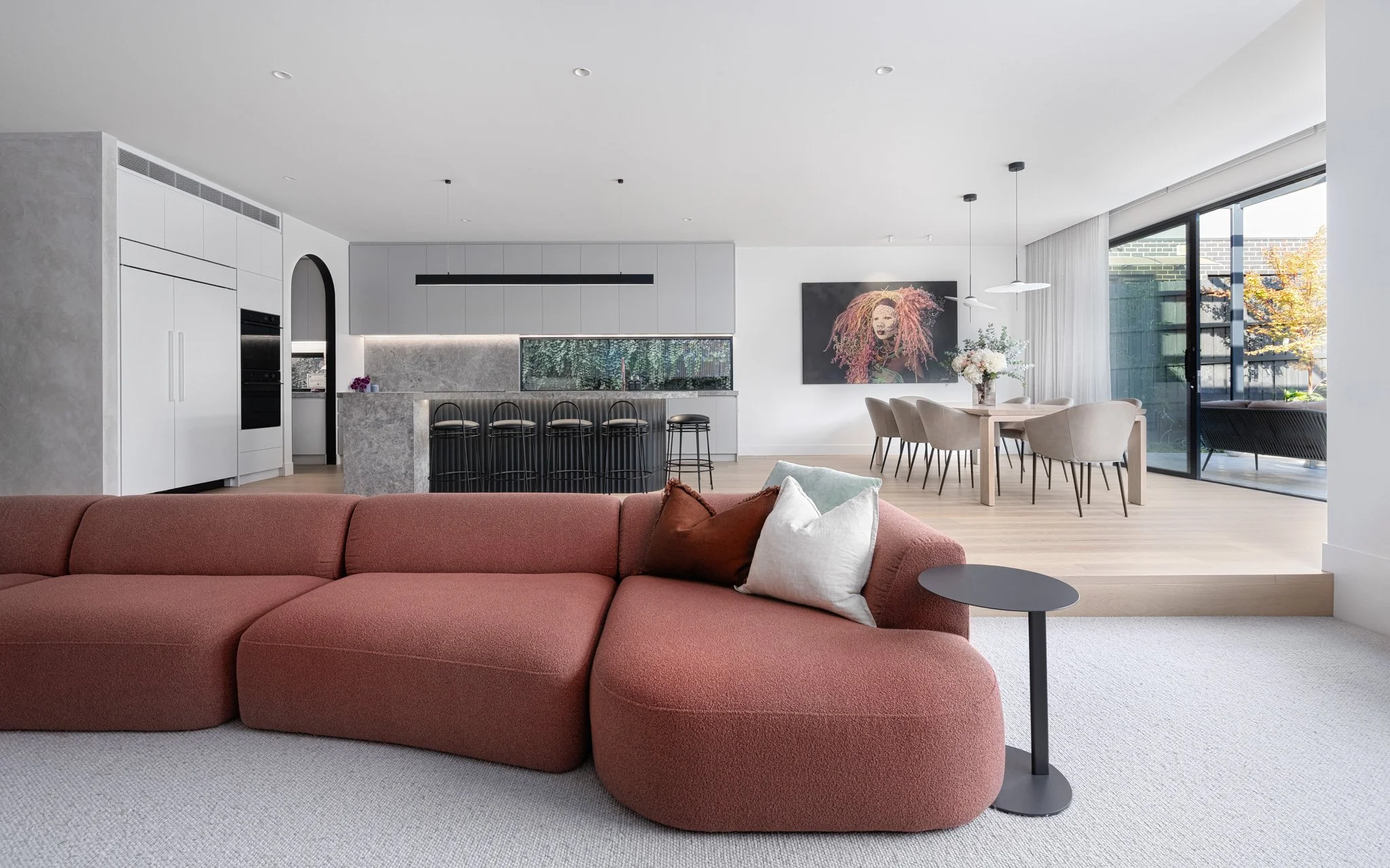 Open-concept living room with pink sectional sofa, black side table, kitchen with bar seating, dining area with beige chairs, large glass sliding doors, and contemporary artwork.