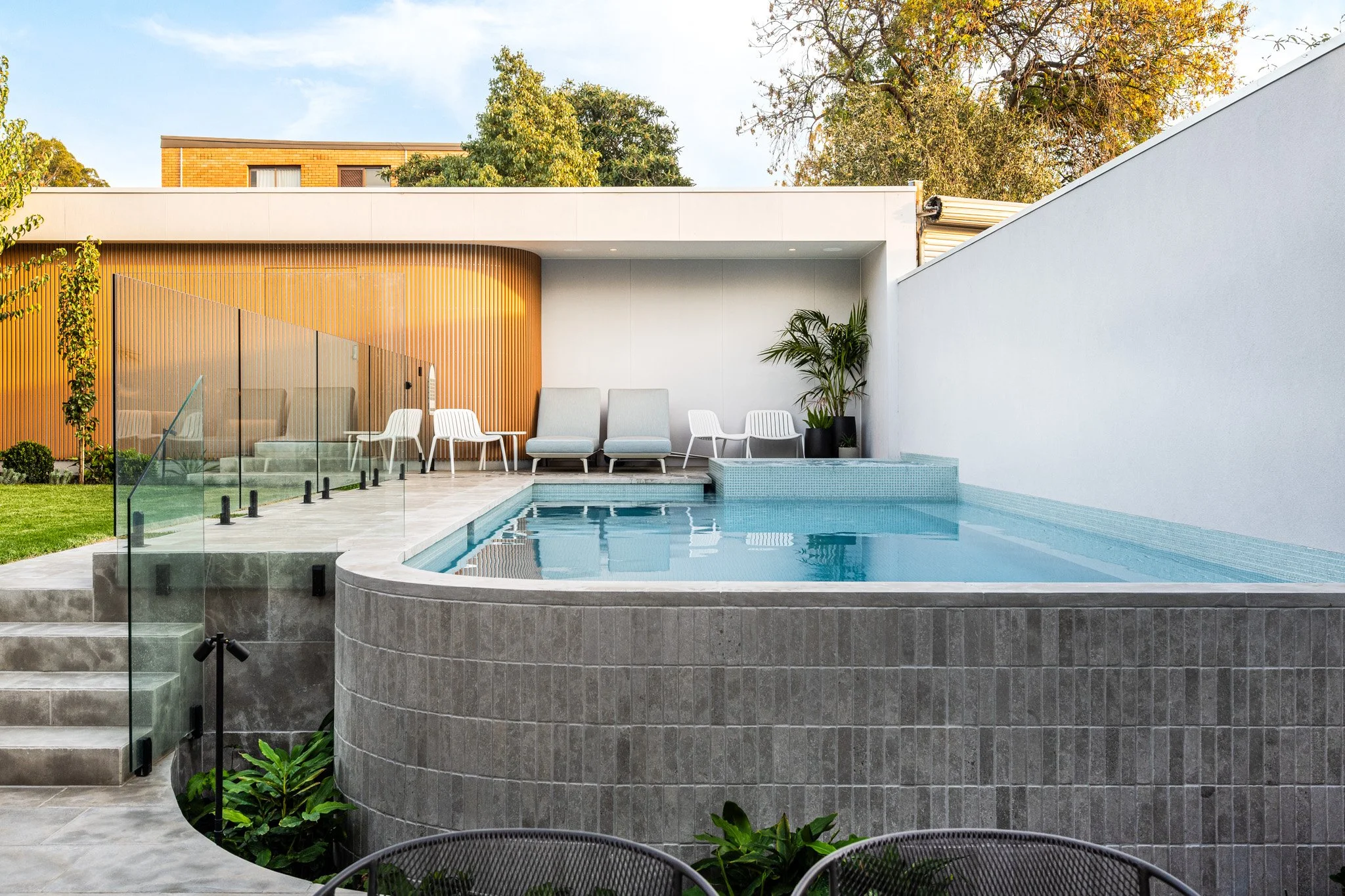Modern backyard with small swimming pool, outdoor seating on patio, potted plants, and a landscaped garden, in sunny weather.