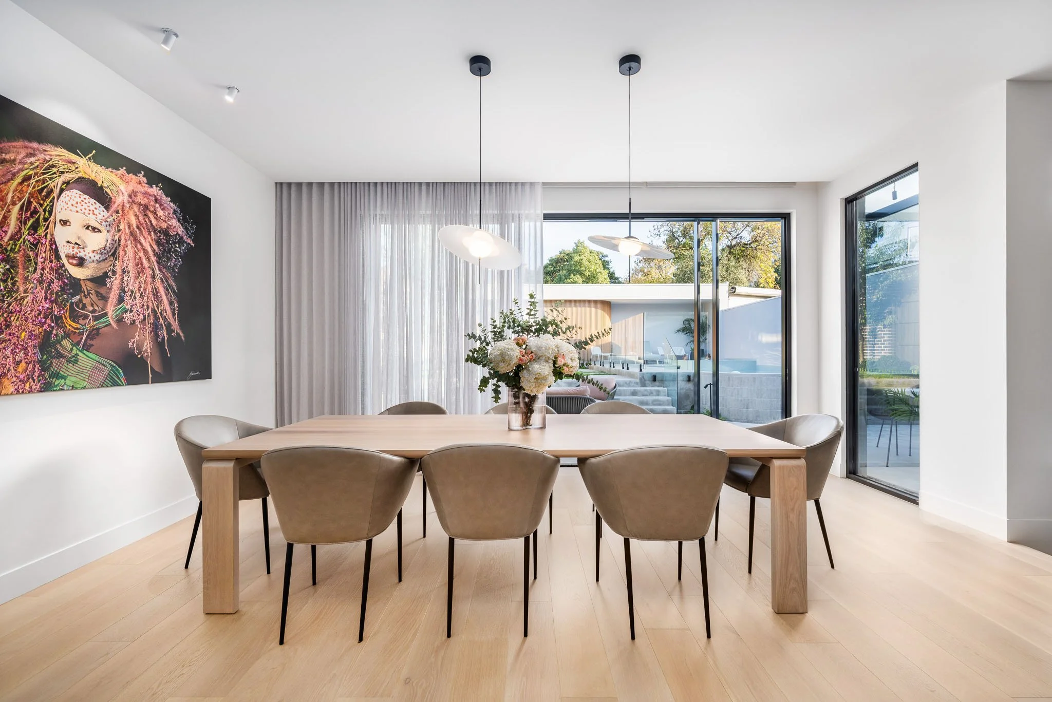 Modern dining room with a light wood table, ten beige chairs, a flower arrangement on the table, large sliding glass doors, and artwork of a woman with colorful hair on the wall.