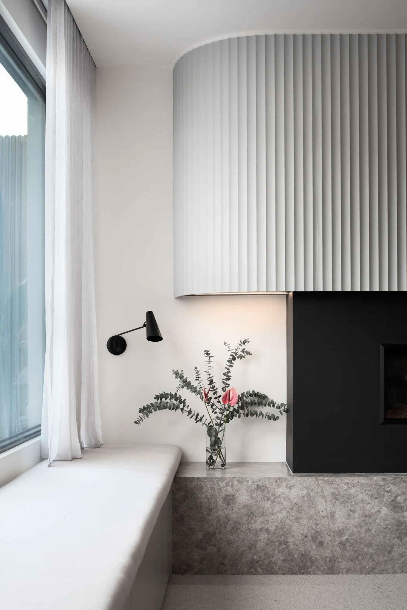 Interior of a modern room with a large window, white curtains, a black wall-mounted reading lamp, a vase with eucalyptus and pink flowers on a stone ledge, and a wall with white and black sections and vertical textured panels.