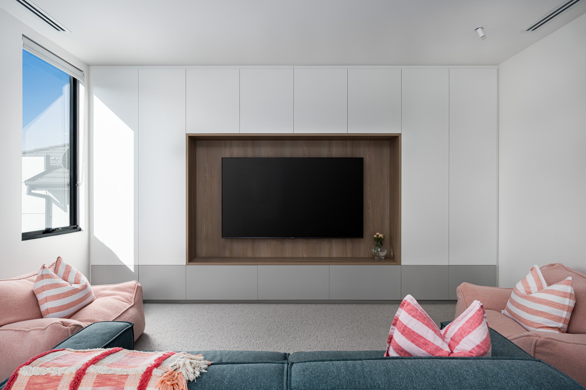 Modern living room with pink sofas, striped pillows, a black TV on a wooden wall panel, and a window with sunlight.