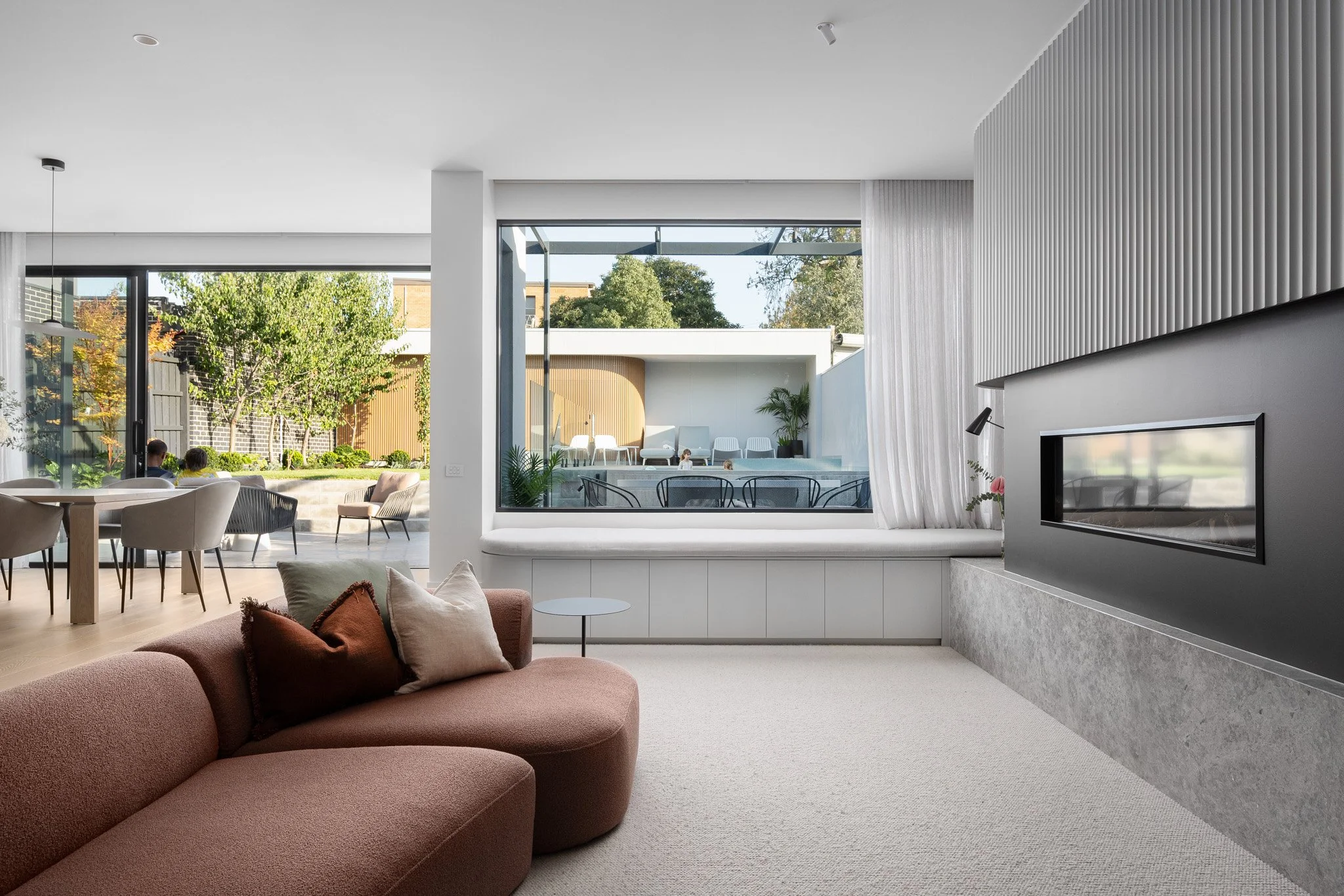 Modern living room with a pink sectional sofa, decorative pillows, glass window seat, gray fireplace, and large windows showing outdoor seating and greenery.