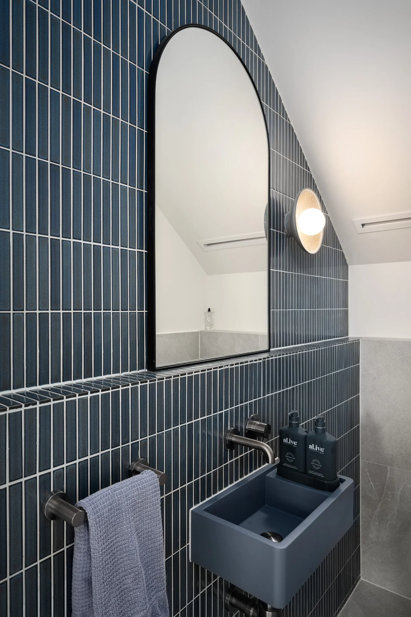 Modern bathroom with dark blue tiled wall, a mirror, a wall light, a small blue sink, and wall-mounted towel and soap dispensers.