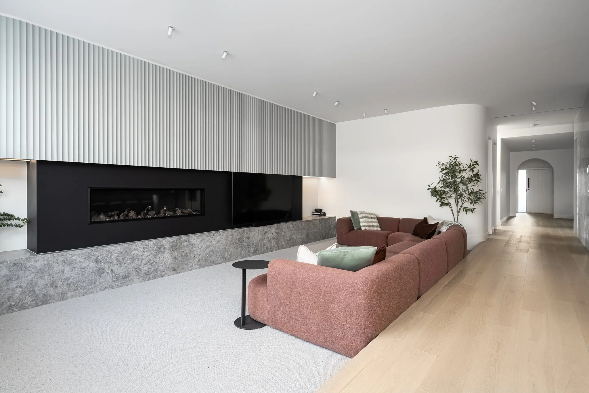 Modern living room with a peach sofa, green and white pillows, a small black side table, a large flat-screen TV, a plant, and a wall-mounted fireplace with a gray marble base.