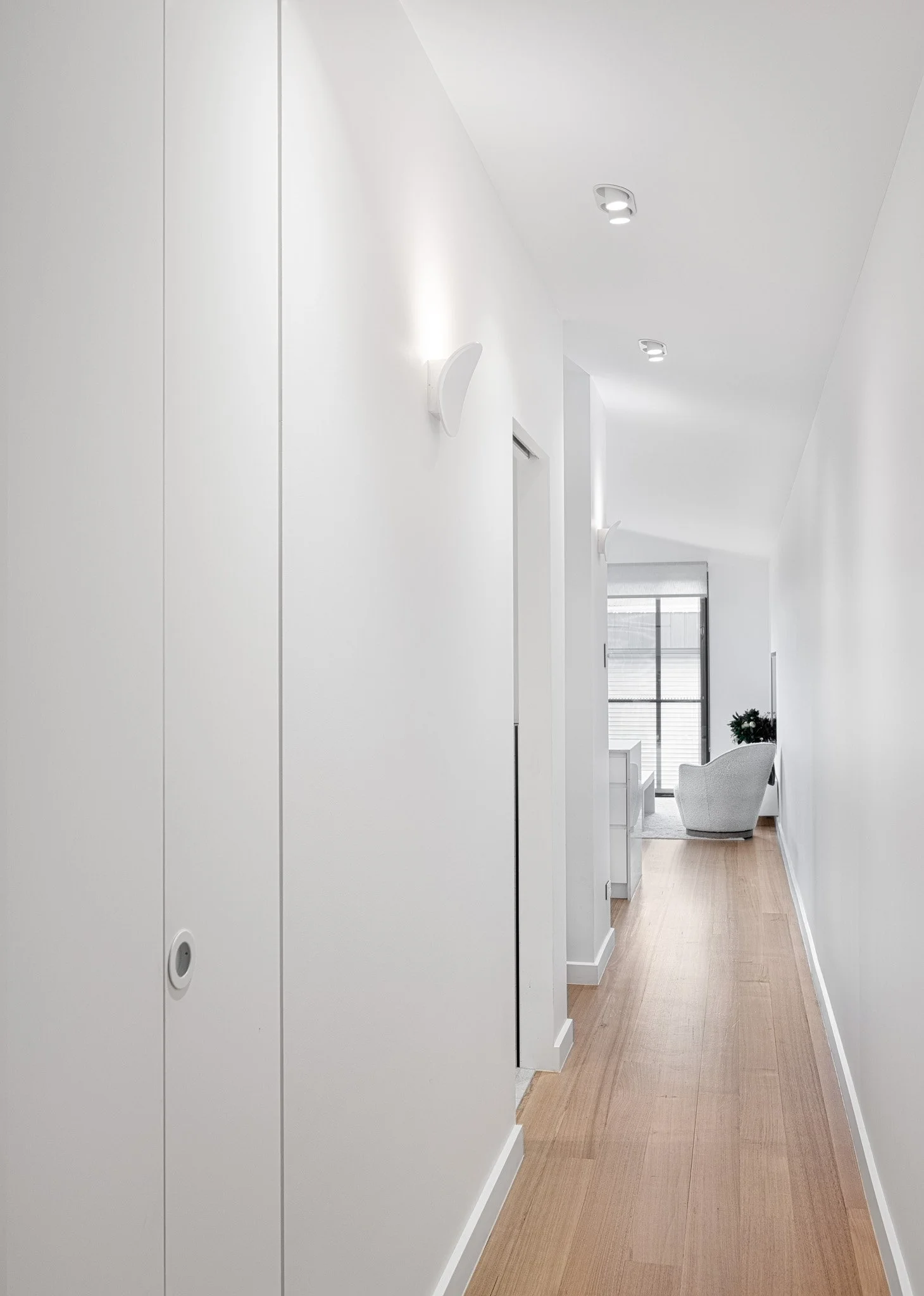 A narrow, minimalist hallway with white walls, wooden flooring, and modern ceiling lights, leading to a sitting area with a white chair and large window.