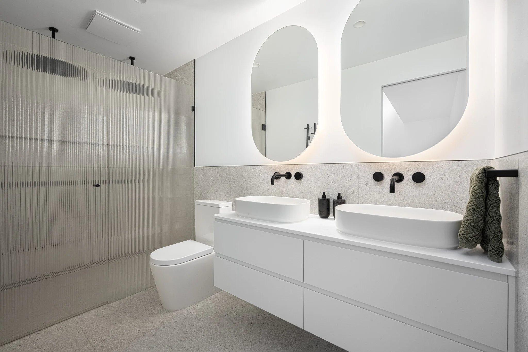 Modern bathroom with large illuminated mirrors, white double sink vanity, black fixtures, gray textured wall, and metallic sliding door.