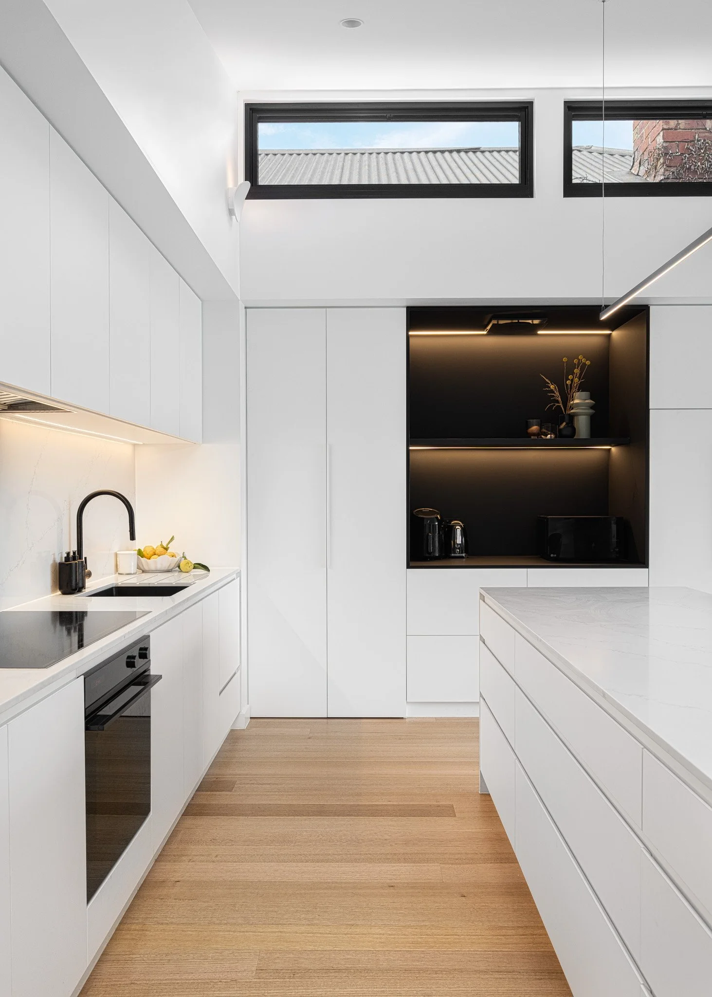 Modern kitchen with white cabinetry, a marble countertop, black appliances, and a black nook with shelves holding small decor items and a few appliances. Large windows near the ceiling with a view of the roof and sky.