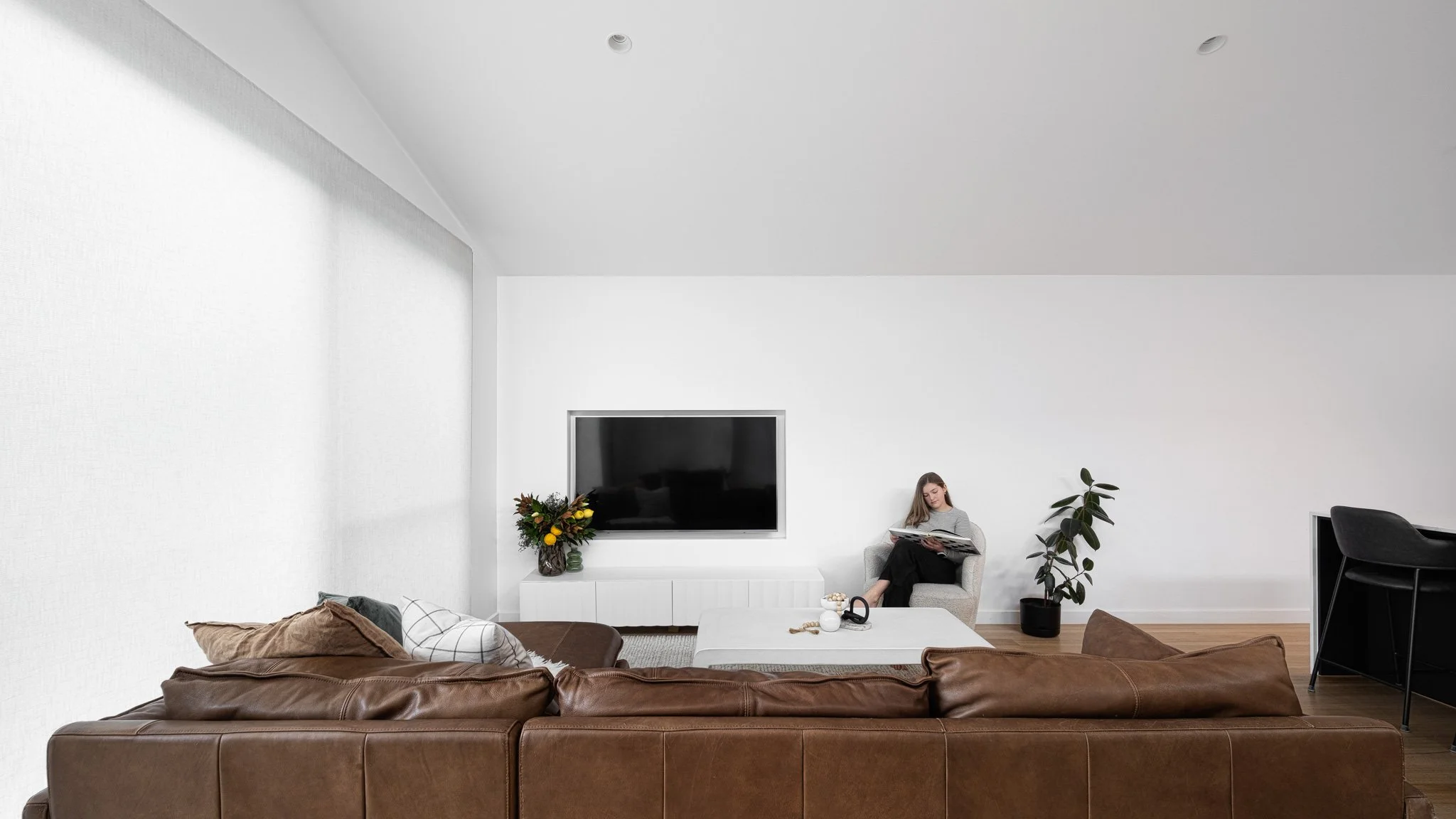 Living room with brown leather sofa, white coffee table, black armchair, TV mounted on white wall, woman sitting on armchair reading a book, potted plant, flowers in a vase, wooden floor, white walls.