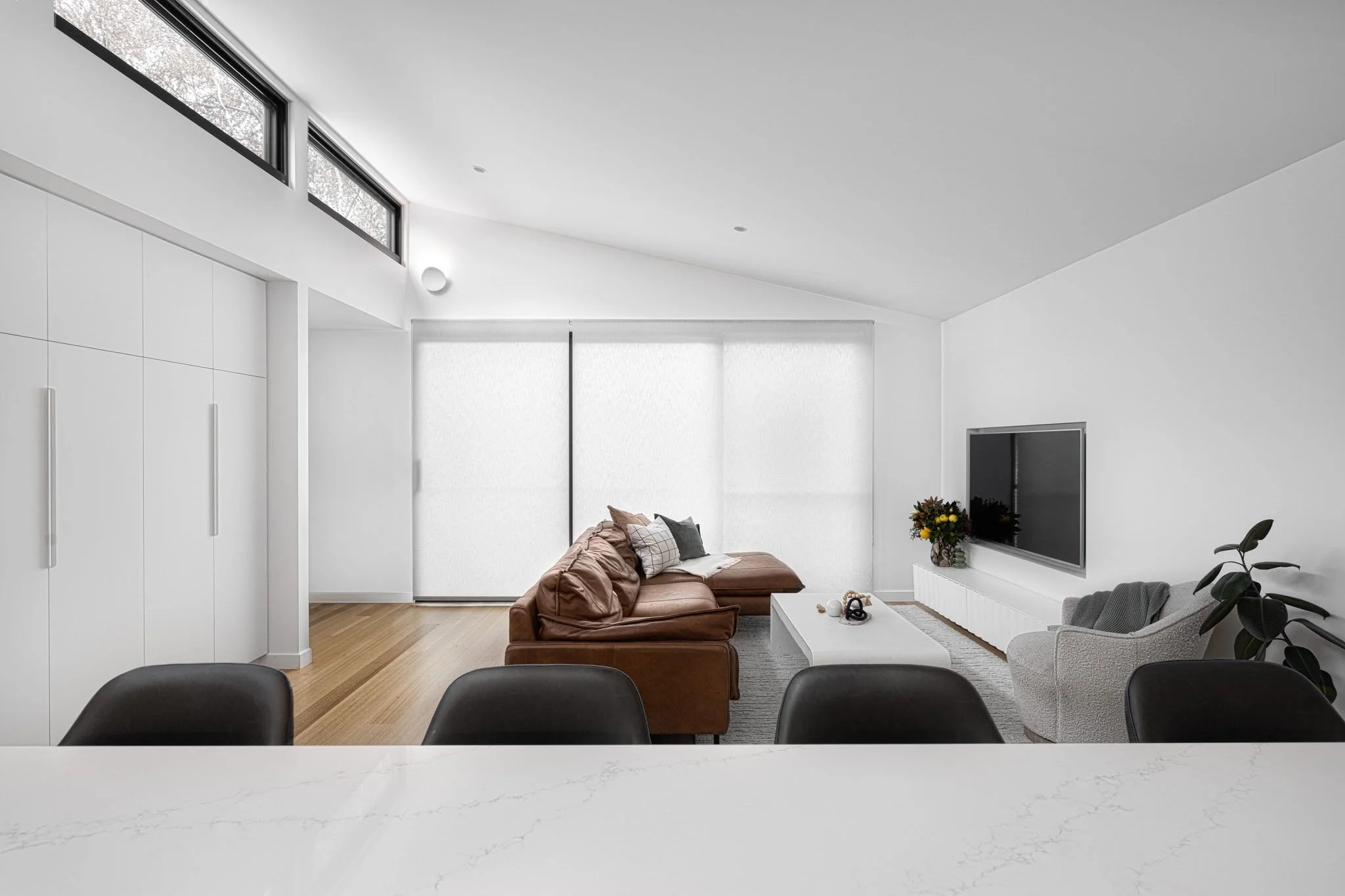 Modern living room with white walls, large windows, a brown leather sectional, a gray armchair, a white coffee table, and a flat-screen TV mounted on the wall.