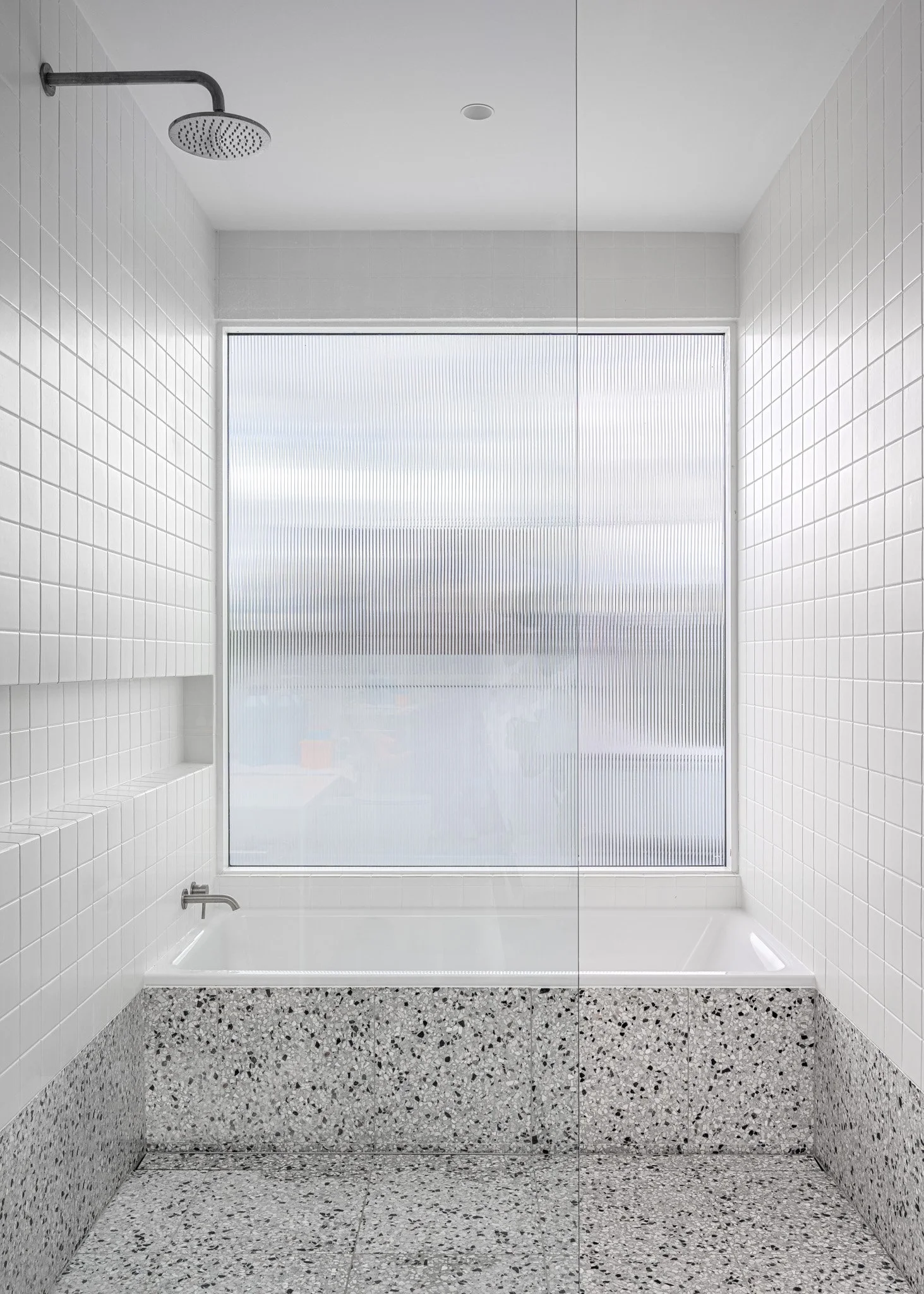 Empty modern bathroom with white tiled walls, terrazzo bathtub, large frosted window, showerhead, and minimalist design.