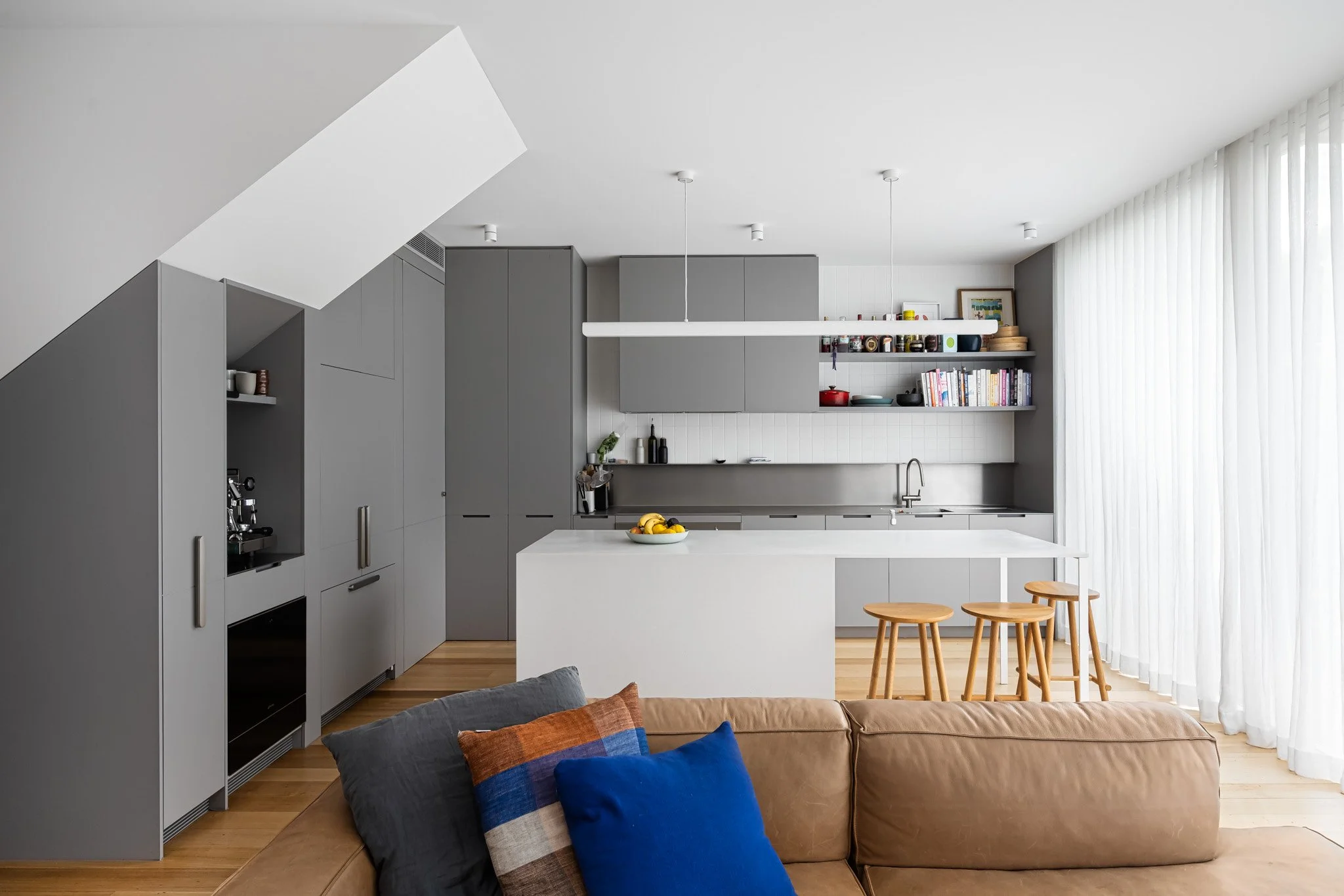 Modern kitchen with grey cabinets and white island, open shelving with books and dishes, beige sofa with colorful pillows in the foreground, large window with white curtains.
