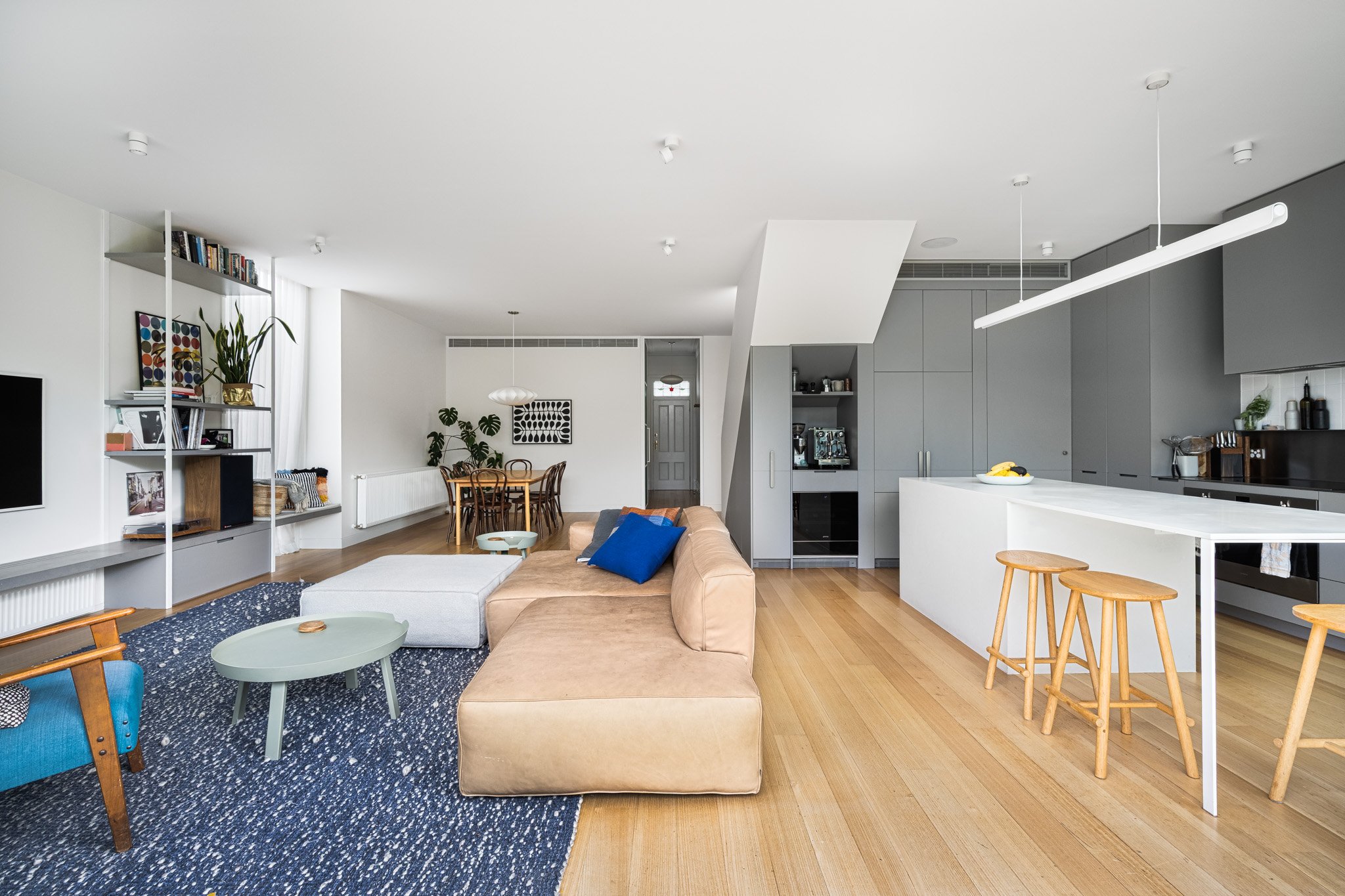 Open-concept living room and kitchen with modern decor, a tan sectional sofa, navy area rug, white walls, wooden flooring, dining table with chairs, and gray cabinetry in the kitchen.