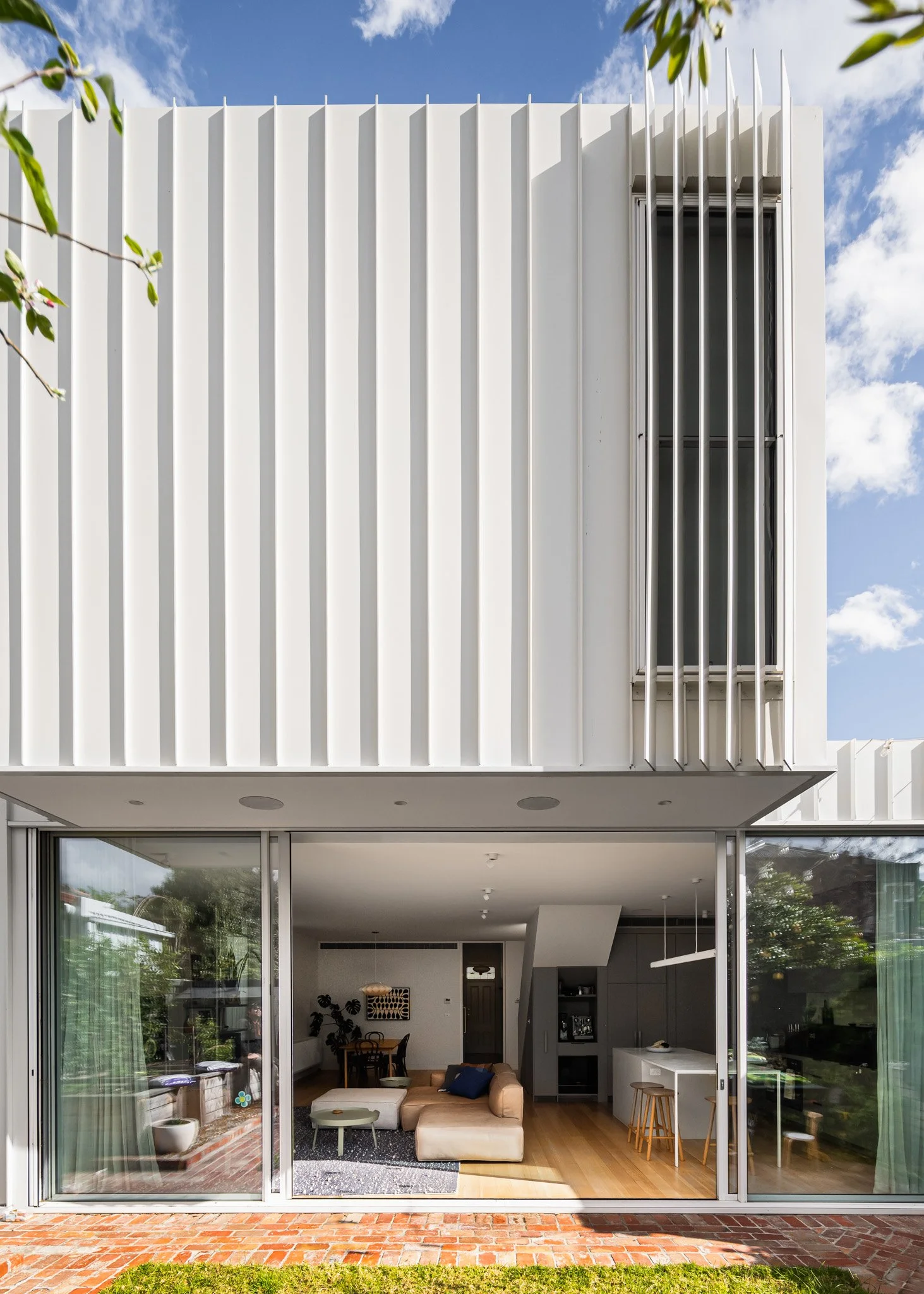 Modern white house with vertical paneling, large sliding glass doors opening to living room with beige sectional sofa and kitchen visible inside, brick patio in foreground, trees and blue sky with clouds outside.