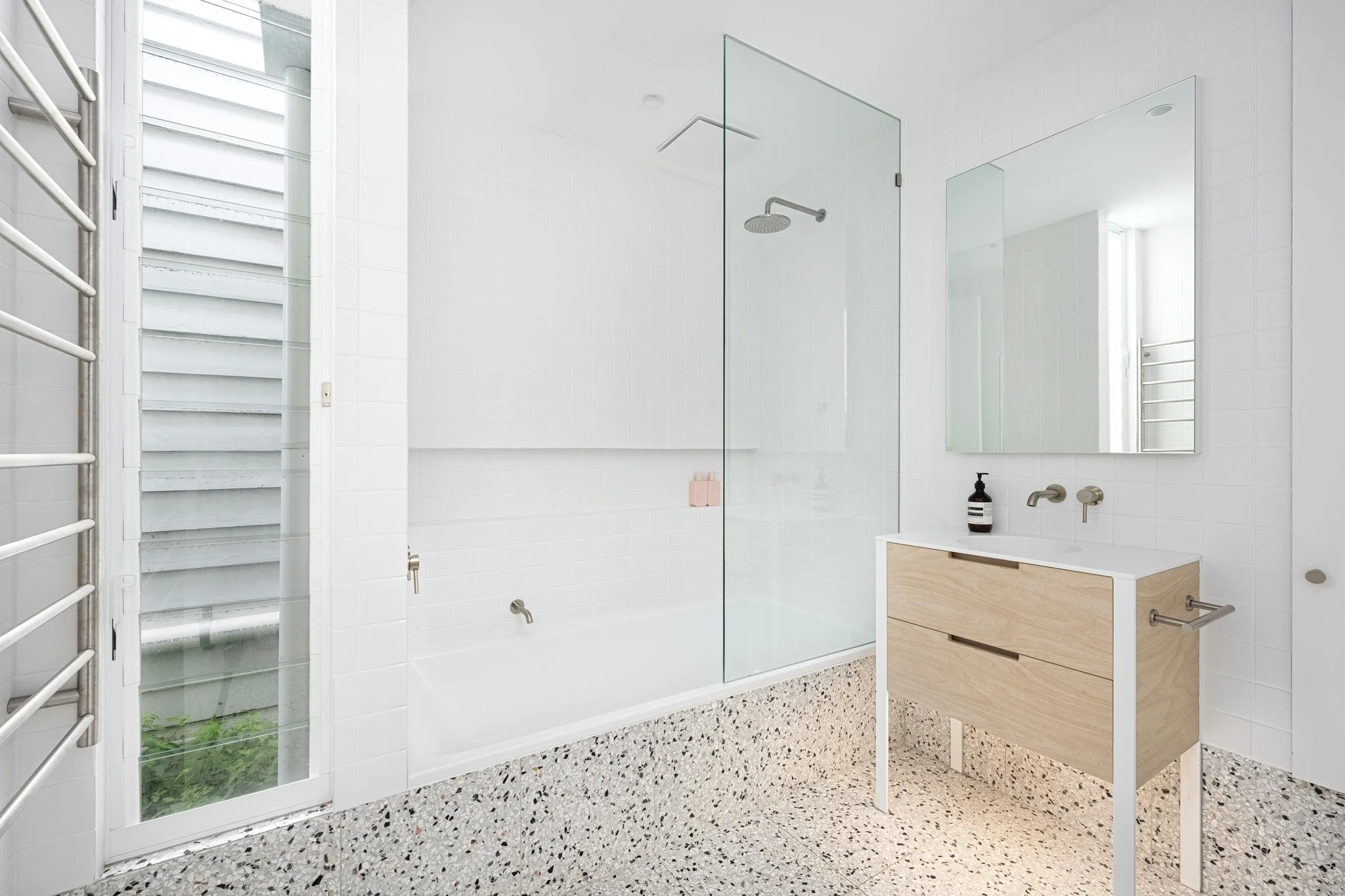 Modern bathroom with white tiled walls, a glass shower enclosure, a freestanding wooden vanity with a white top, a large mirror, a black soap dispenser, and a reflective window with a heated towel rail.