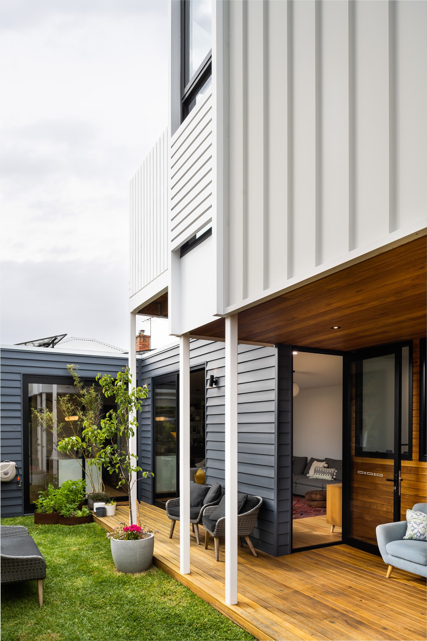 Modern house with black and white siding and a wooden porch, outdoor seating area with chairs, potted plants, and a grassy yard.