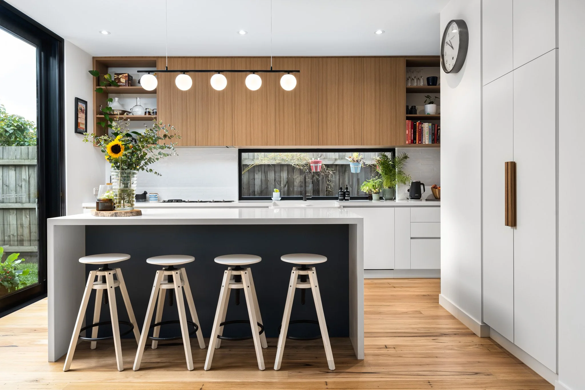 Modern kitchen with white cabinets, a black-topped island, four white bar stools, wooden flooring, and a window with outdoor greenery.