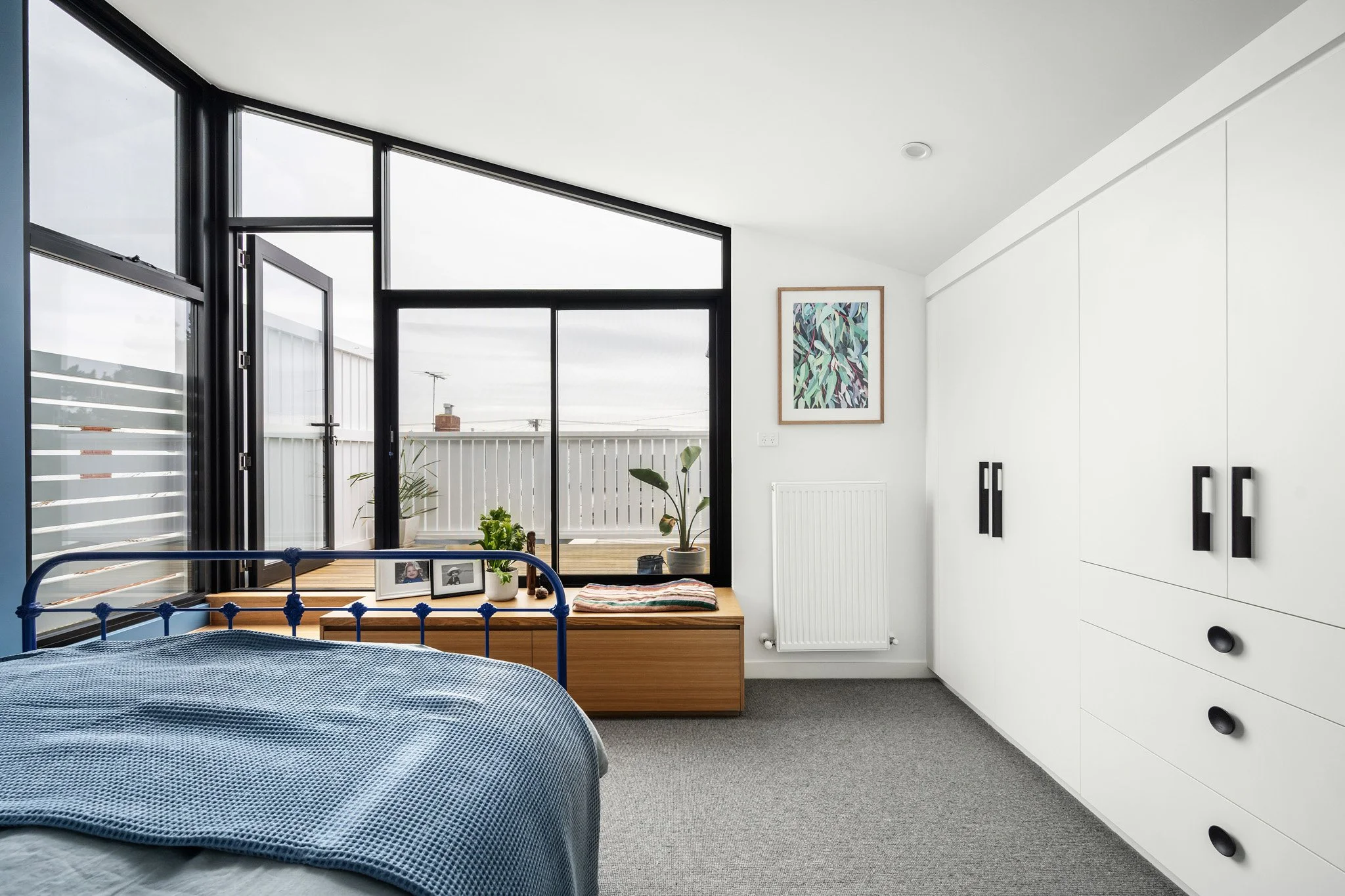 Modern bedroom with large black-framed windows, white walls, gray carpet, white built-in wardrobe with black handles, a bed with blue bedding, a wooden bench with plants and framed photos, and framed artwork on the wall.