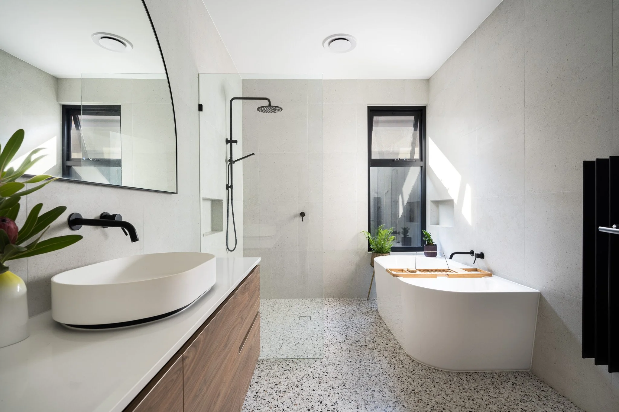 Modern bathroom with a floating vanity, vessel sink, black faucet, wall-mounted showerhead, a bathtub with plants nearby, and a large mirror, illuminated by natural light from a window.
