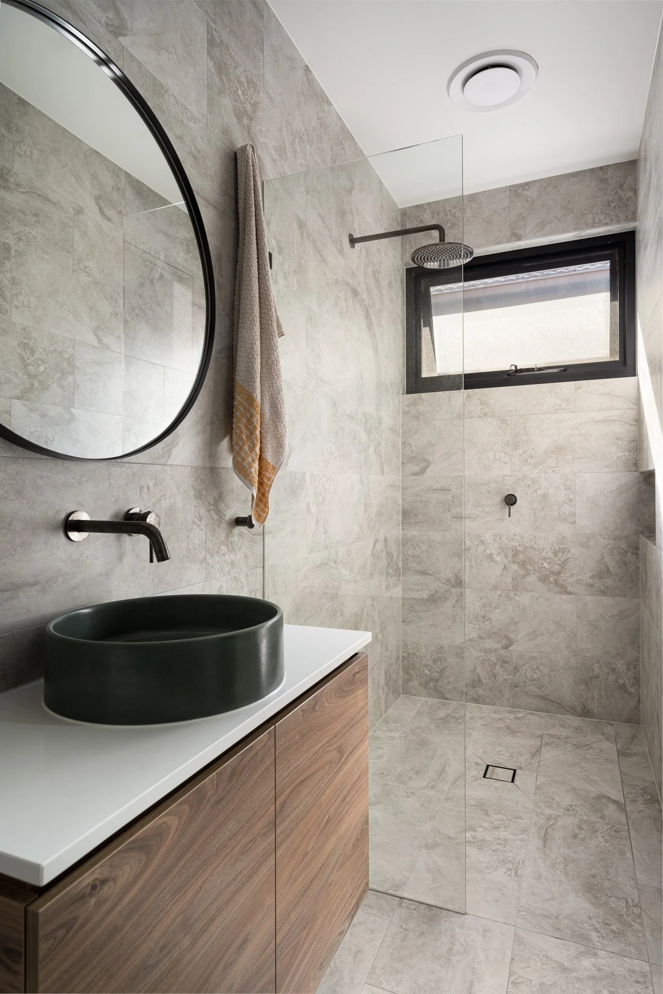 Modern bathroom with a round mirror, beige tiled walls and floor, black sink, wooden vanity, towel hanging on the wall, black-framed window, rain shower head, and a small drain on the floor.