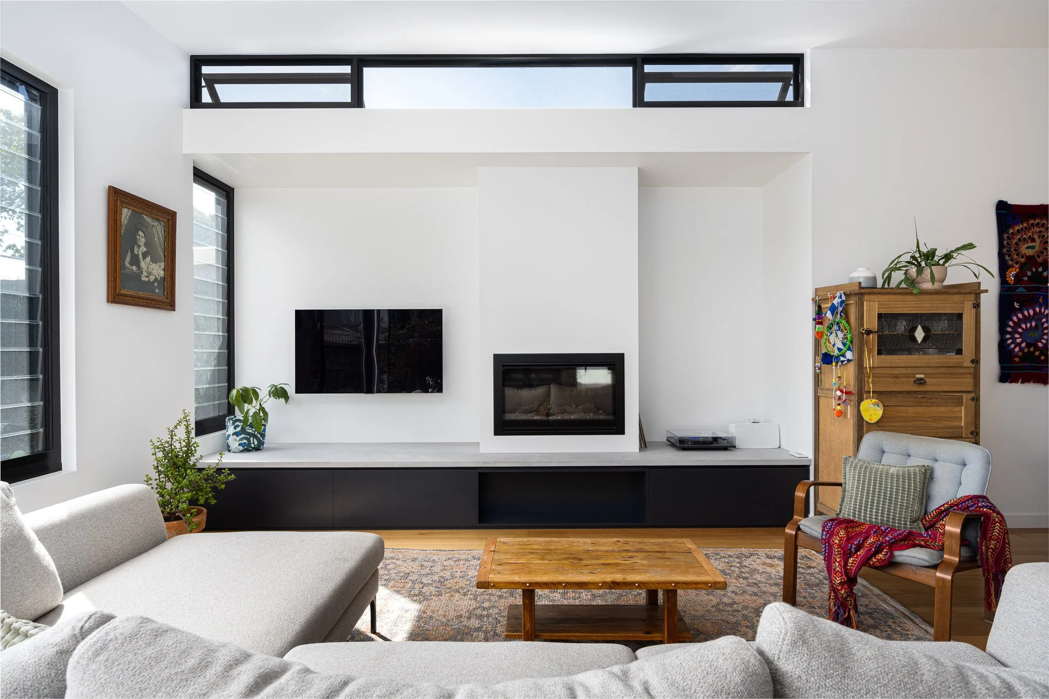 Modern living room with white walls, large windows, wall-mounted TV, fireplace, wooden furniture, potted plants, and cozy seating area.