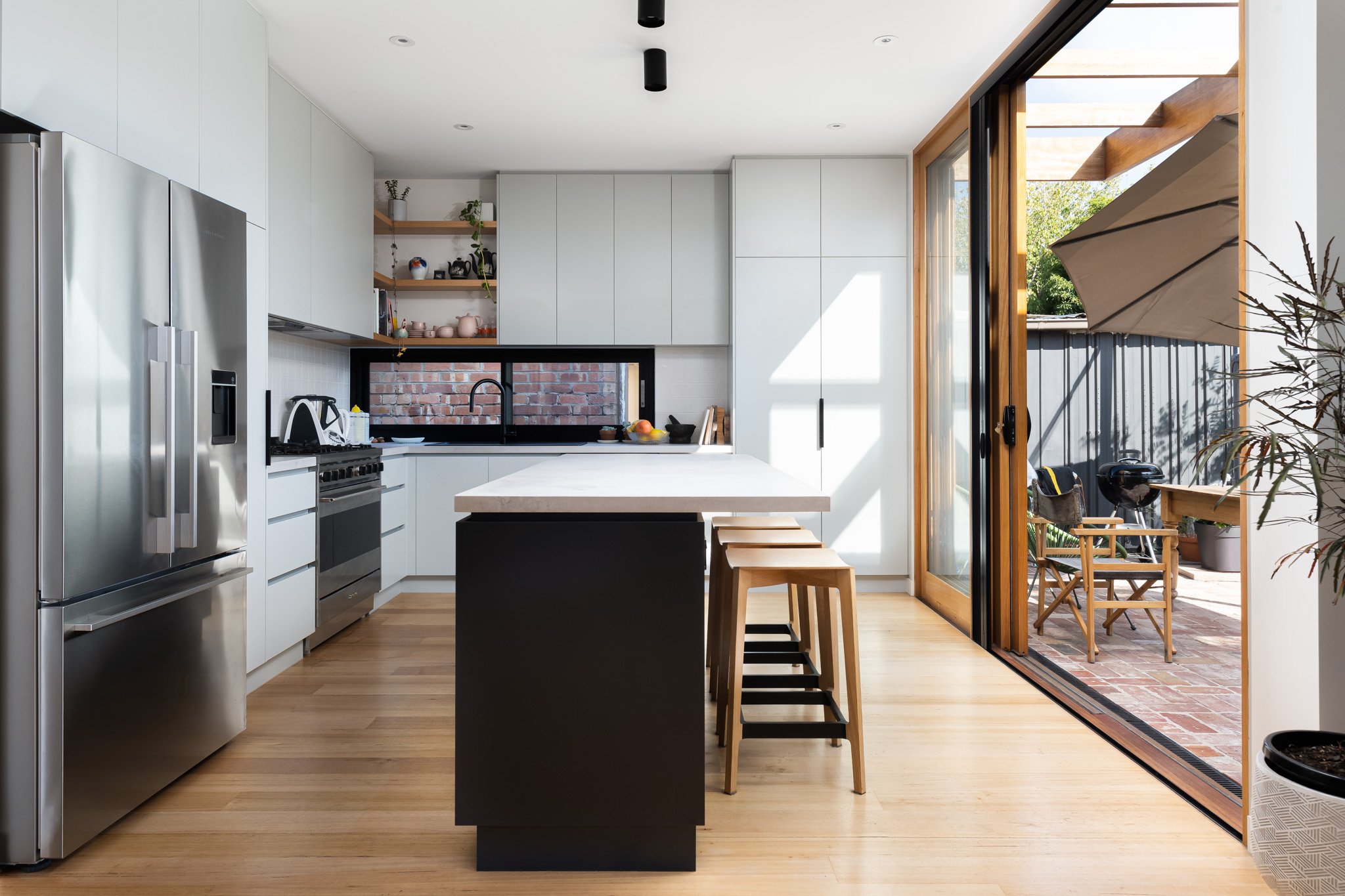 Modern kitchen with white cabinets, stainless steel appliances, black countertop, and a black-framed window. An island with a white countertop and four wooden barstools is in the center. Sliding glass door opens to a patio with chairs, an umbrella, a