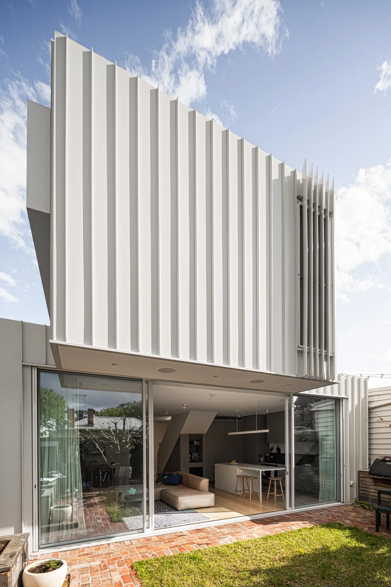 Modern white two-story house with vertical siding and large sliding glass doors opening to a backyard patio, green grass, and brick paving.