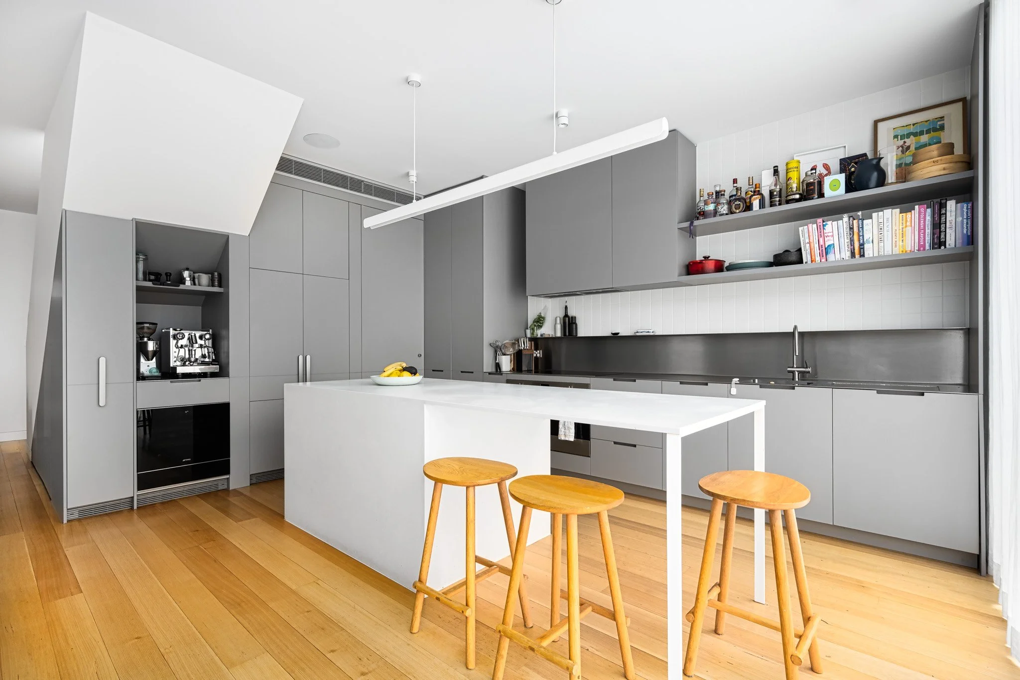 Modern kitchen with gray cabinets, white countertop island, wooden flooring, open shelves with cookbooks and bottles, minimal decor, three wooden stools, and a window with curtains.