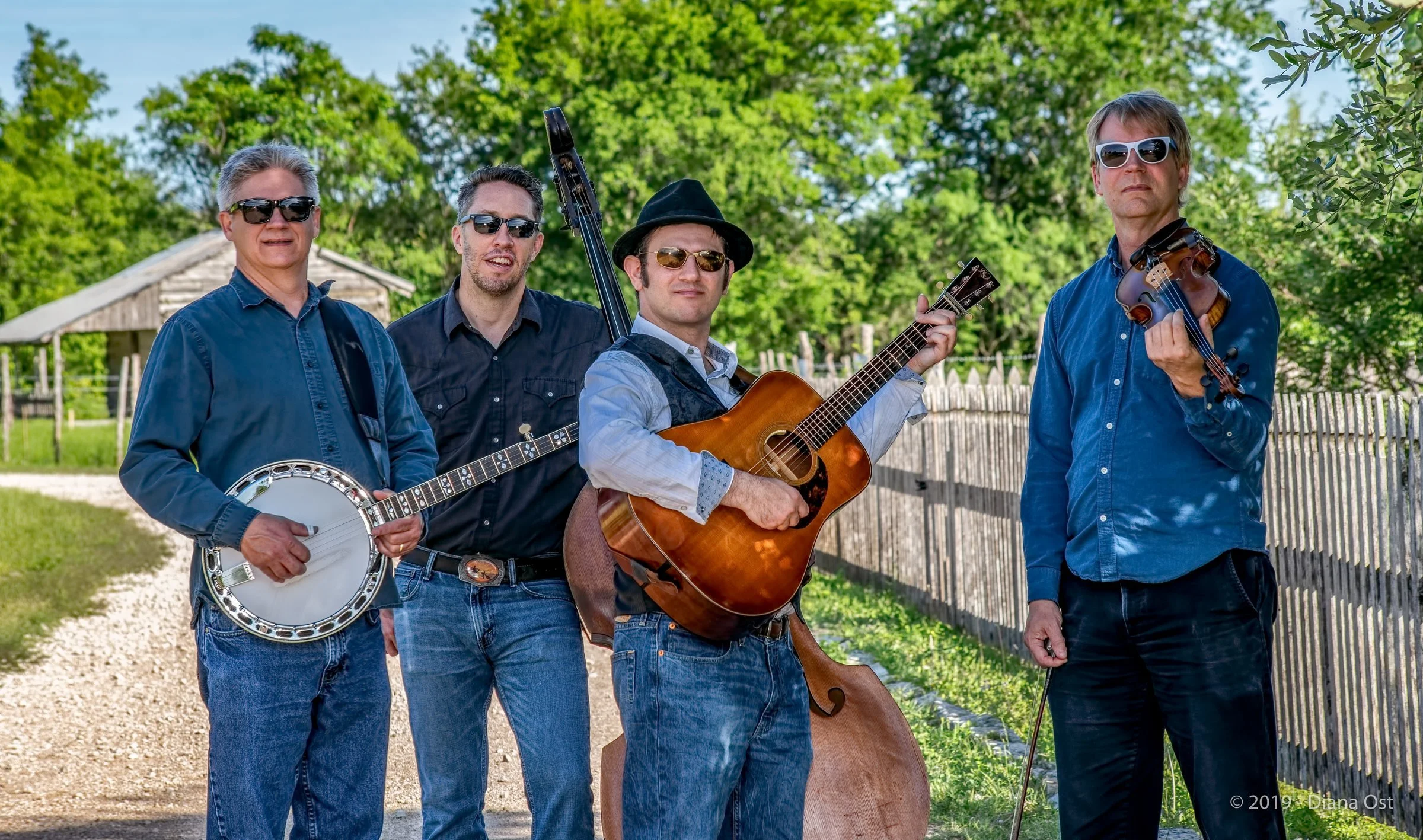 Hot Pickin 57s bluegrass quartet standing on a gravel path at Pioneer Farms with cedar fence and Texas hill country landscape