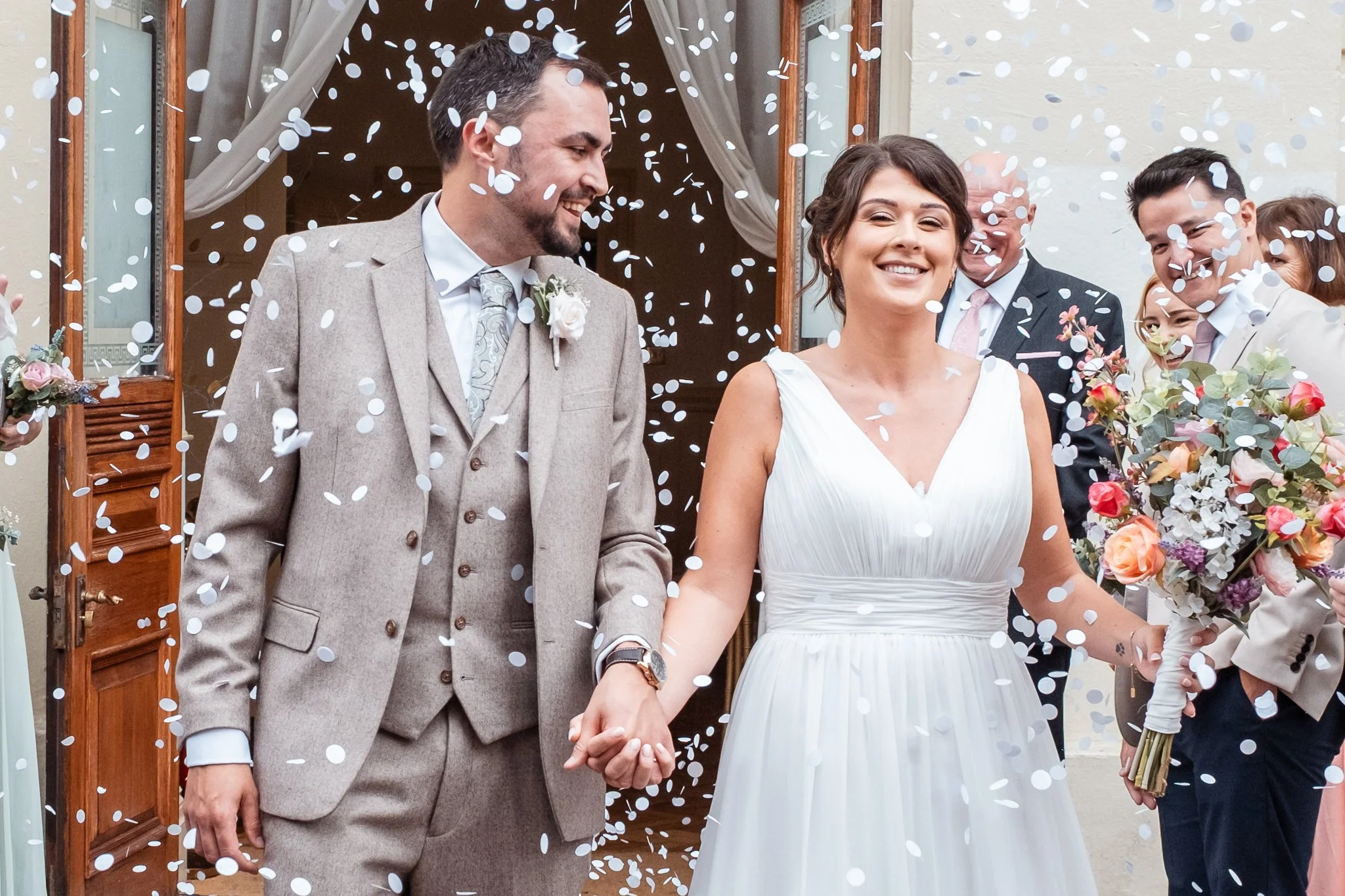 Bride and Groom Holding hands walking through confetti. Smiling. Groom, in light grey three piece suit, looking at Bride adoringly. Bride Holding bouquet