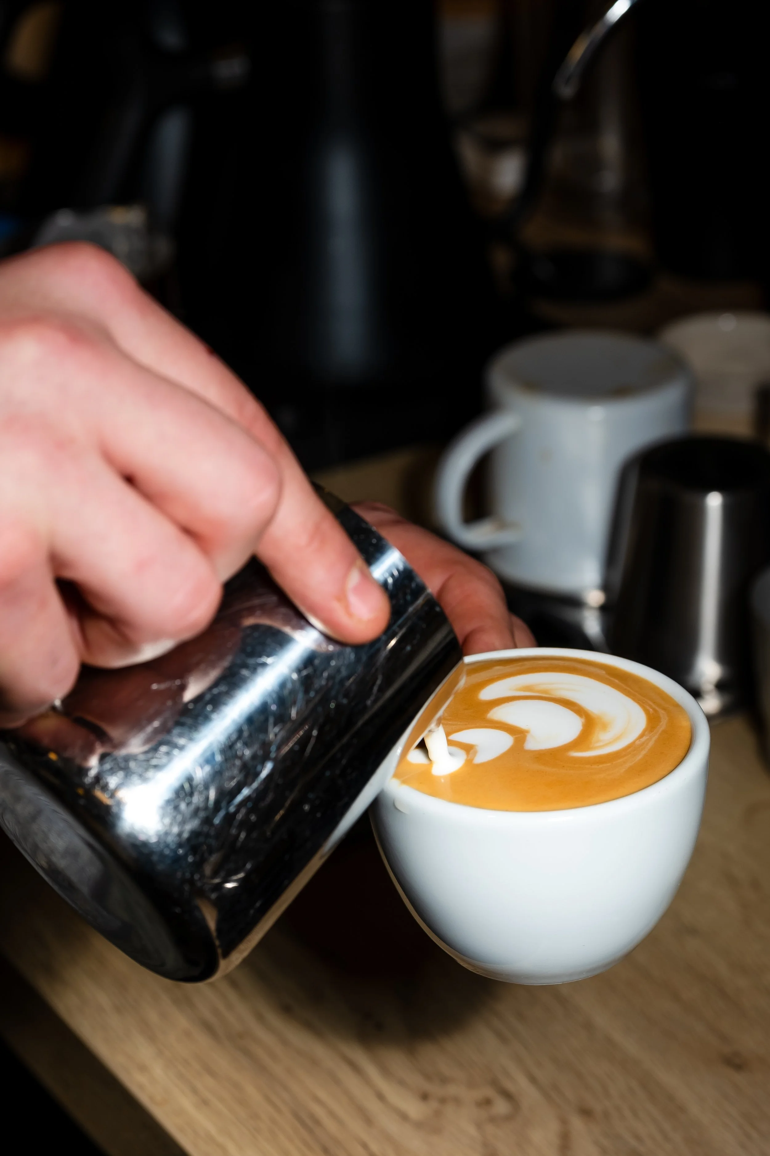 A person pours steamed milk from a metal pitcher into a cup of espresso to make a latte, with latte art on top. In the background, there are various coffee mugs and equipment.