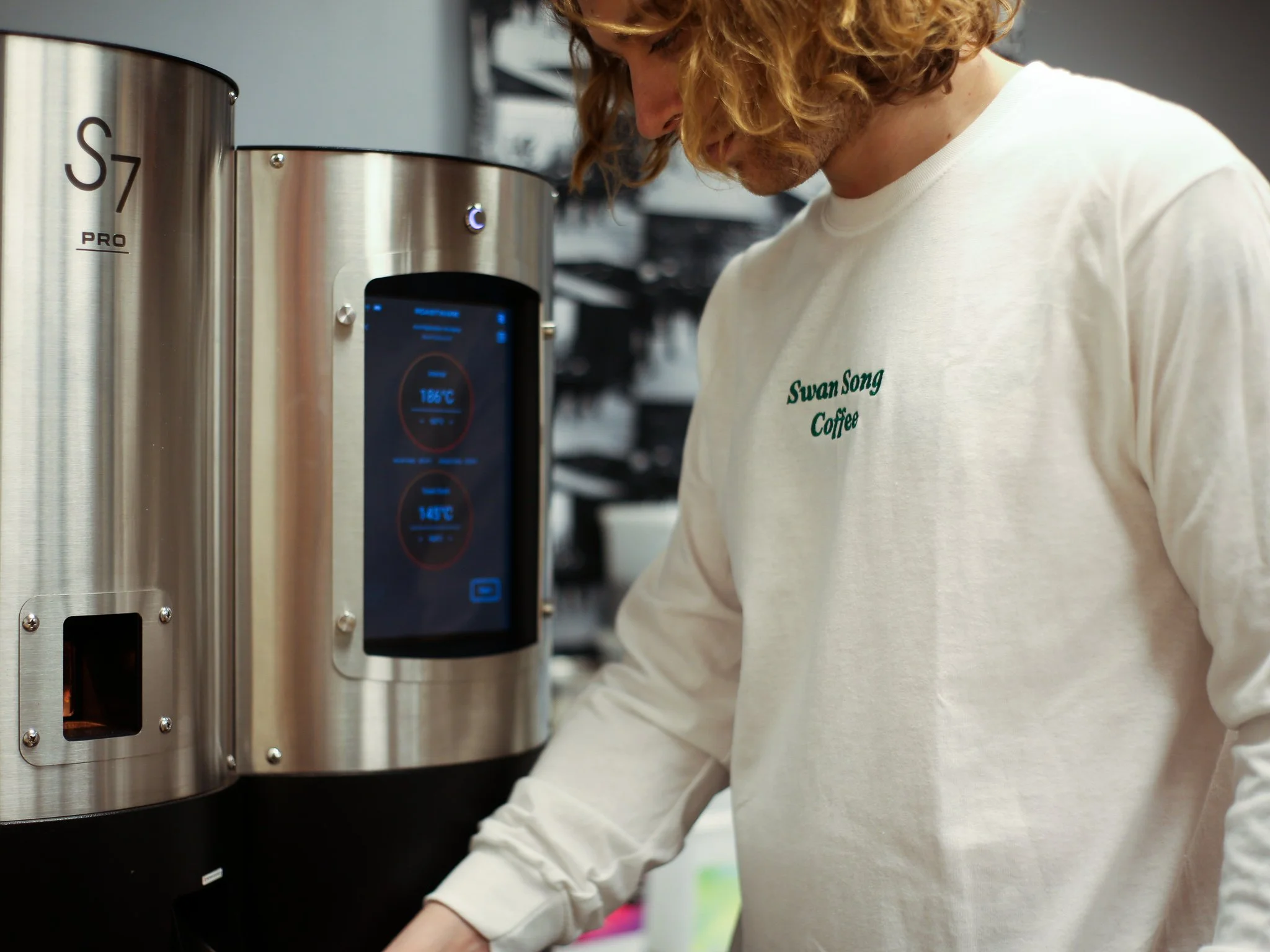 A person with curly hair wearing a white shirt with green embroidery that reads 'Swan Song Coffee' is using a large, stainless steel coffee roaster machine with a digital display showing temperature and other settings.