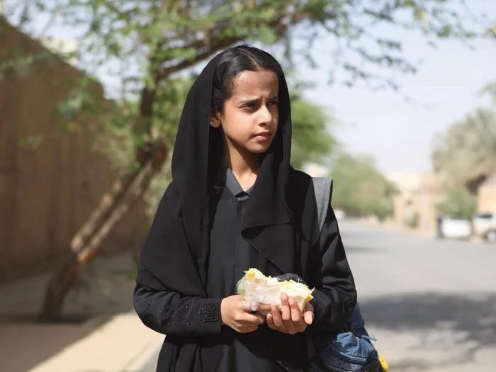 Image description: A girl around 11 years old (Wadjda) stands in the middle of the street holding food in her hands. She is wearing a headscarf.