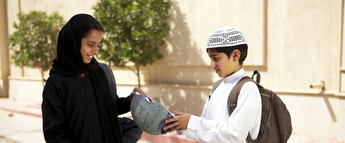 Image description: Wadjda is on the left of the frame in a black headscarf and a black dress holding a backpack. She is being handed a bicycle helmet by the boy on the right in a white shirt, white hat, and holding a brown backpack.