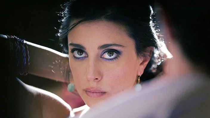 Image description: Layle in Caramel looking at the camera, She has on multiple thin bracelets, jade earrings, and purple eyeshadow.
