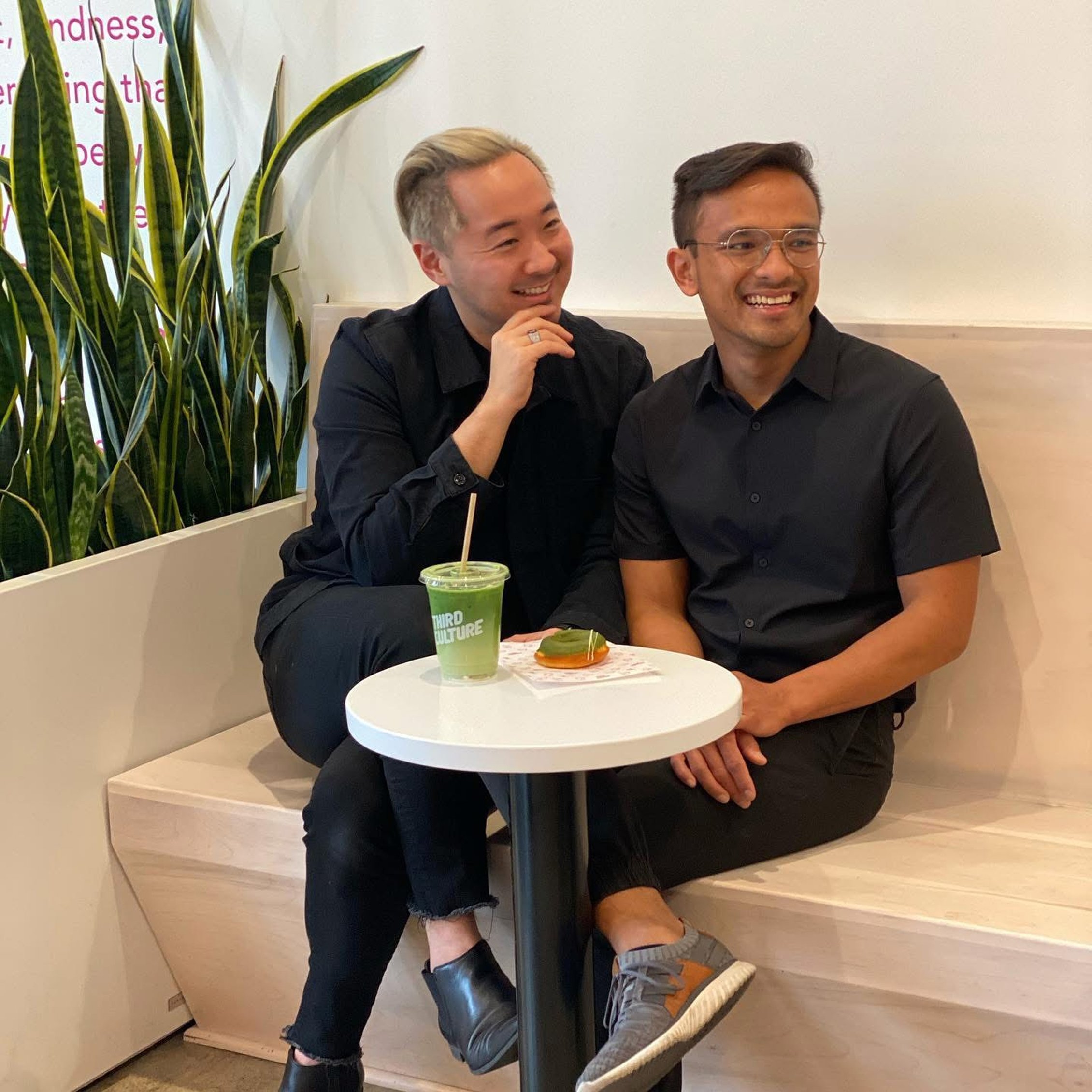 Two men wearing black shirts sitting at a small white table in a cafe, smiling and enjoying each other's company. There is a green drinkand a cookie on the table.