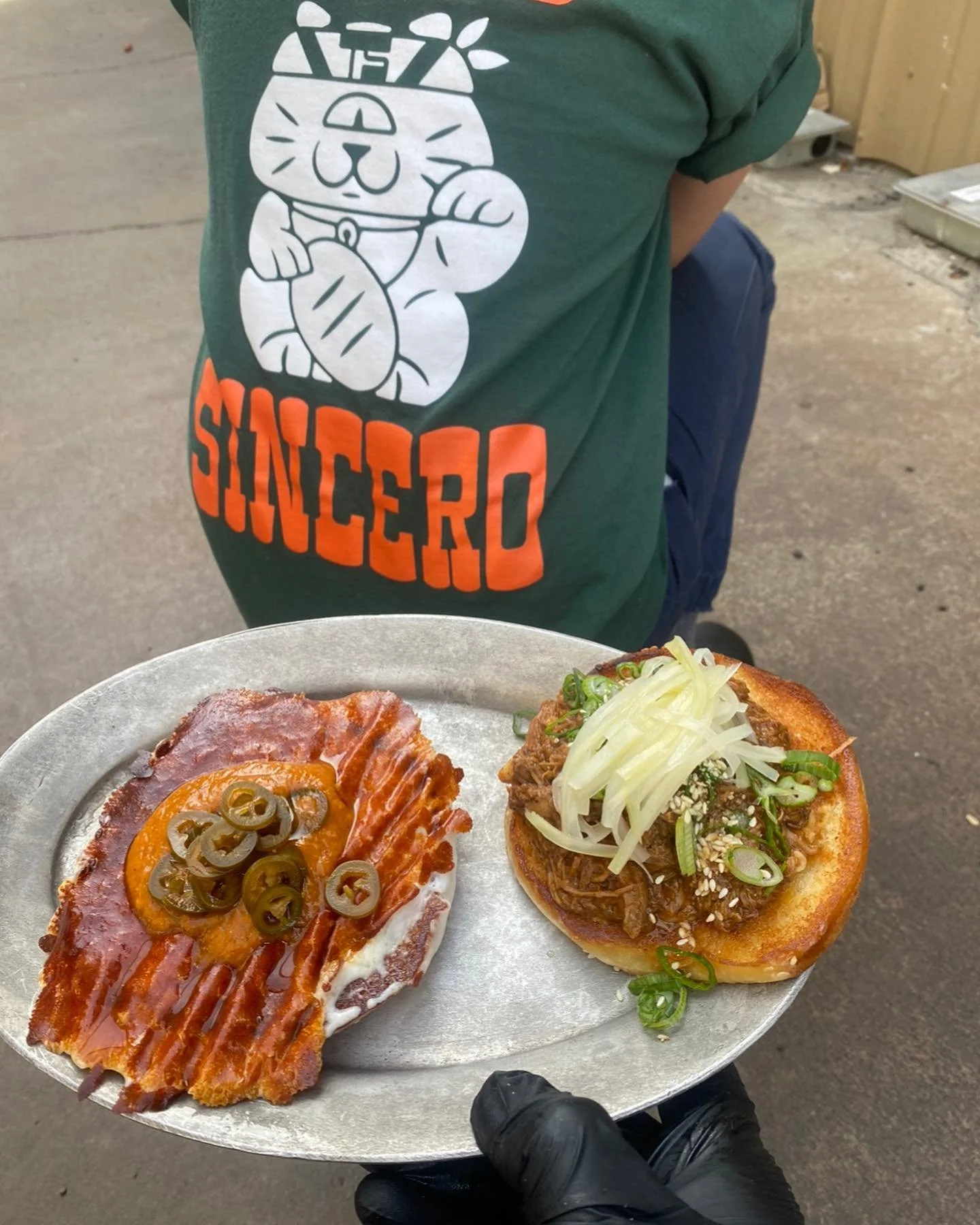 A tray with a bacon and jalapeño slice and a pulled pork sandwich topped with shredded cheese, green onions, and cole slaw, held by a person wearing a green shirt with a cartoon dog and the word 'SHINCERD' written on it.