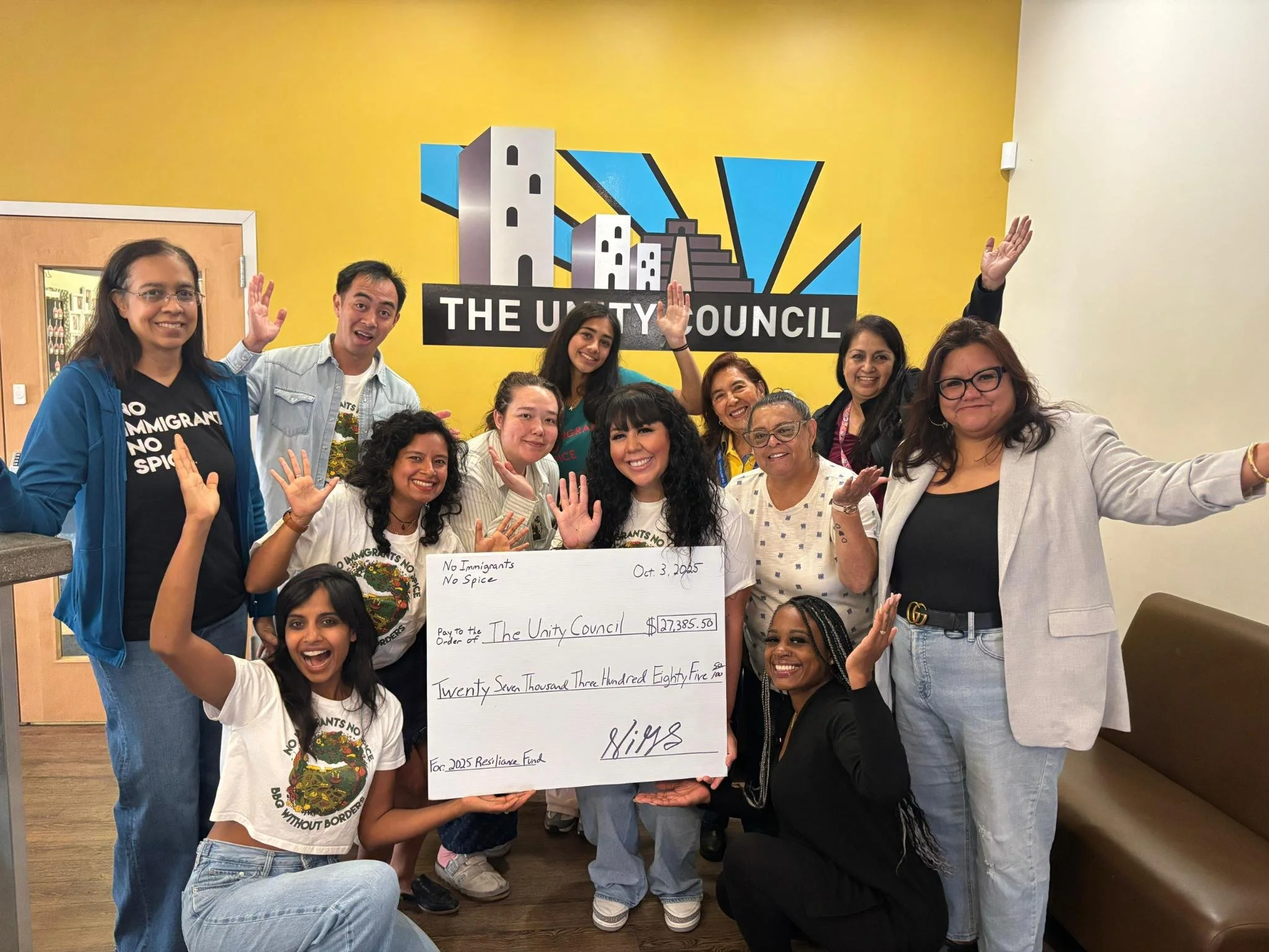 Group of diverse individuals celebrating, holding a large check for $27,385.50, in front of a yellow wall with a cityscape logo and the words 'The Unity Council'.