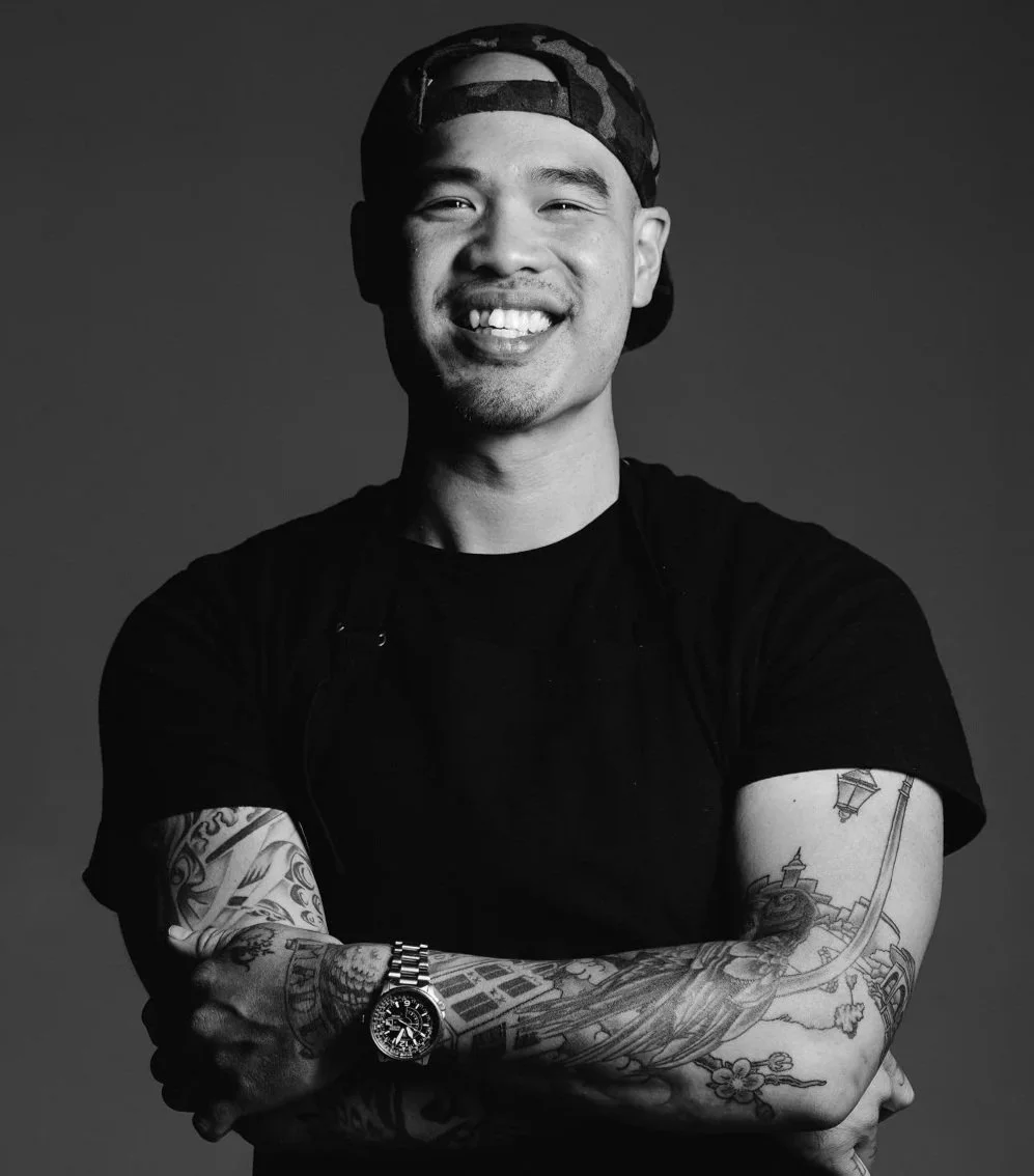 A smiling man with tattoos on his arms, wearing a black t-shirt, a camouflage cap backwards, and a watch, standing with arms crossed against a gray background.