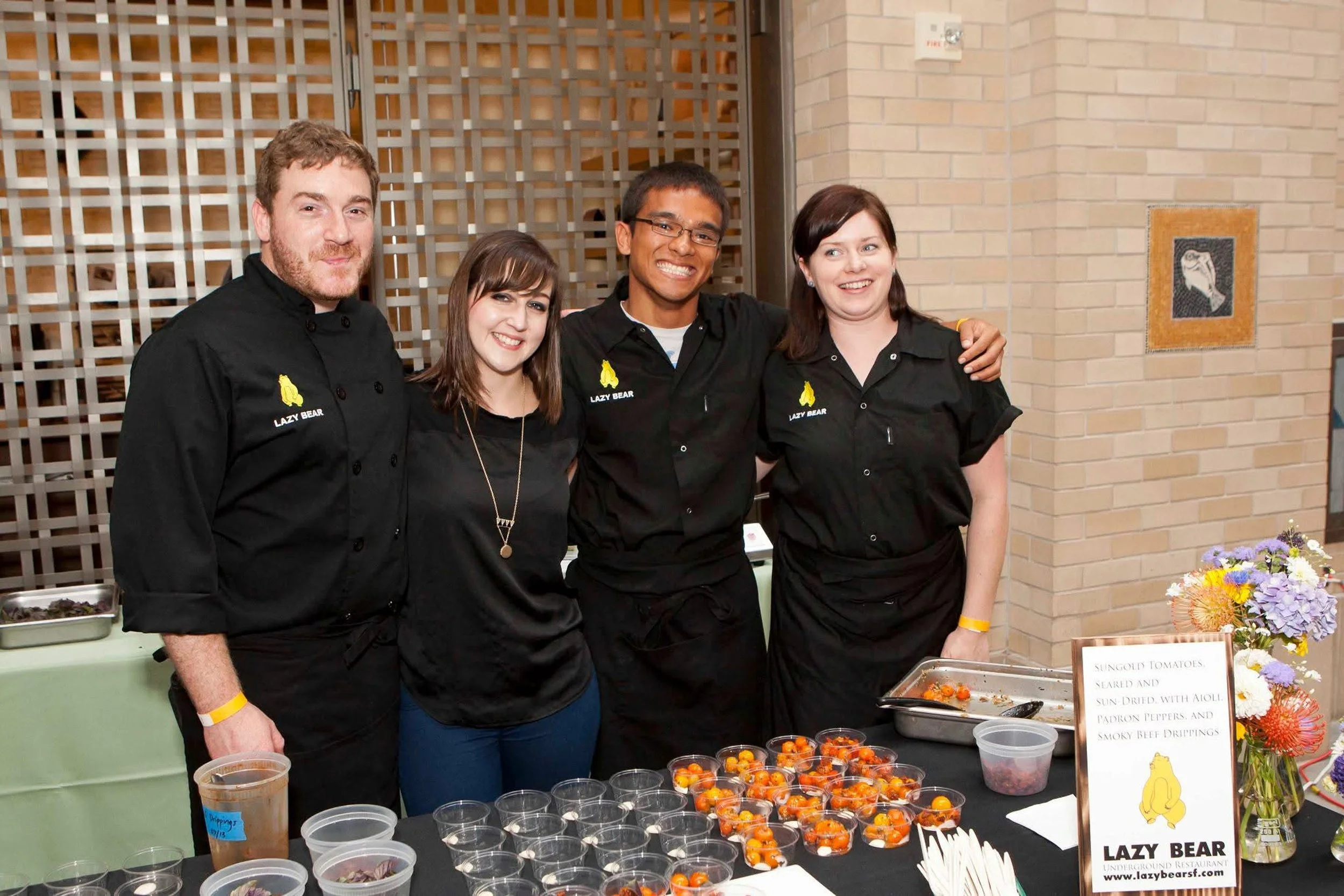 Four people wearing black shirts with a yellow bear logo stand behind a food table at an event. They are smiling and posing with arms around each other.