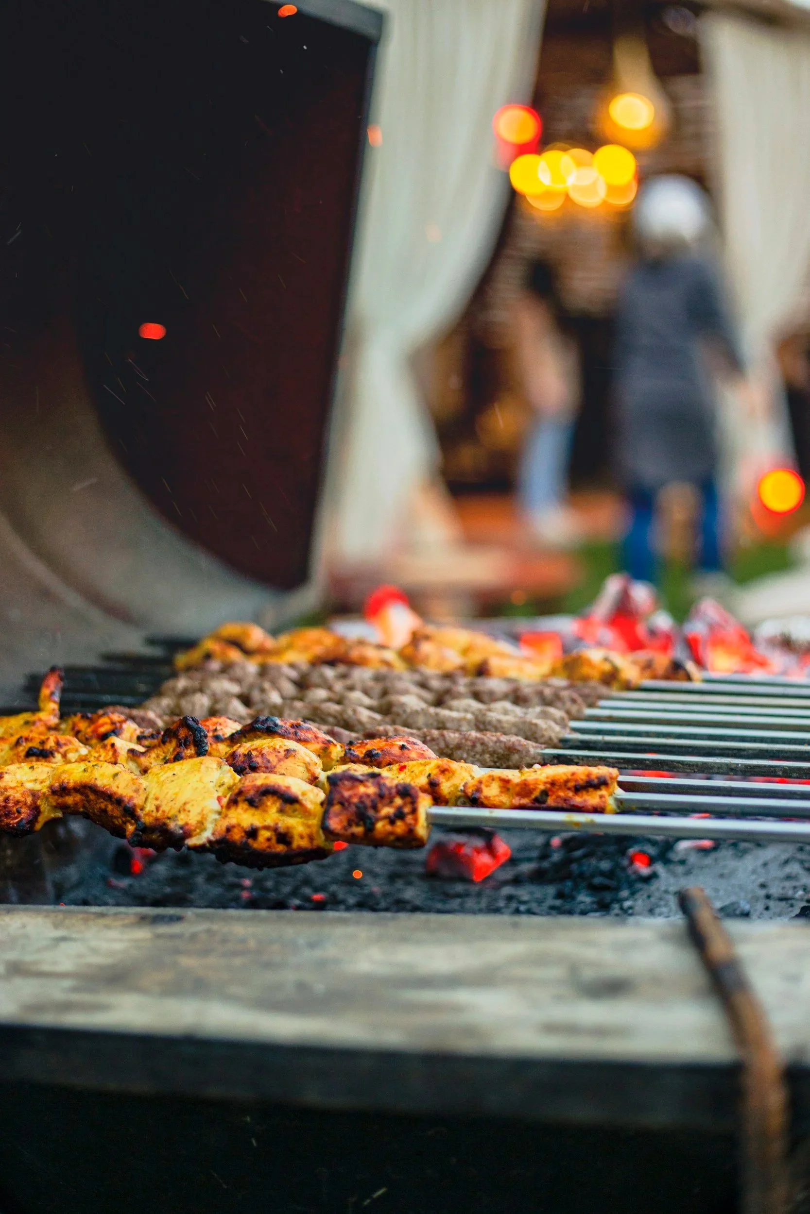Close-up of meat and vegetable skewers grilling over hot coals at an outdoor barbecue, with blurred people in the background.