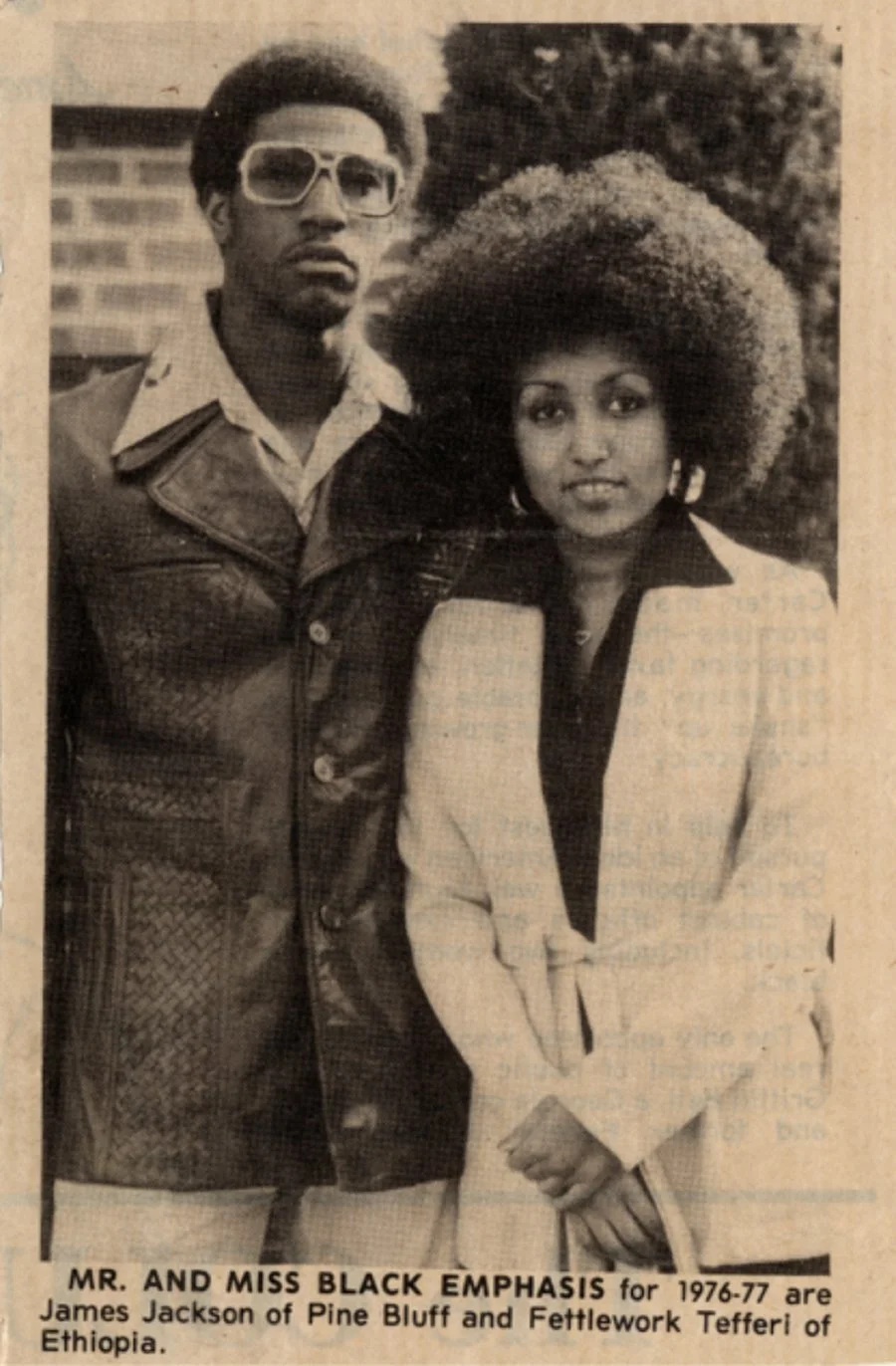 A black-and-white photo of a man and a woman standing outdoors. The man is wearing large sunglasses, a collared shirt, and a leather jacket, and has an Afro hairstyle. The woman has a large Afro hairstyle and is wearing a light-colored blazer over a dark shirt. There are trees and a building in the background. The caption reads:"Mr. and Miss Black Emphasis for 1976-77 are James Jackson of Pine Bluff and Fettlework Tefferi of Ethiopia."
