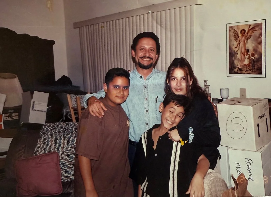 A family of four posing indoors. Two children and two adults, all smiling. The background has a painting of an angel and some moving boxes.