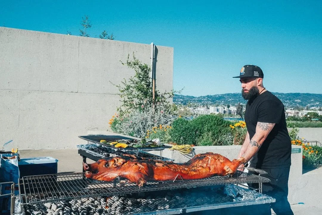 A man in a black t-shirt, with tattoos on his arms, wearing a black cap, is grilling a whole roasted pig on a barbecue outdoors during the day.