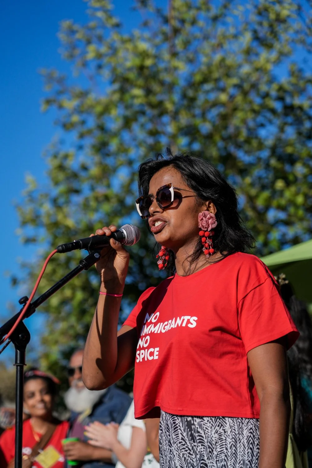woman in red shirt reading "no immigrants no spice" speaking into a microphone with a blue sky and tree in the background