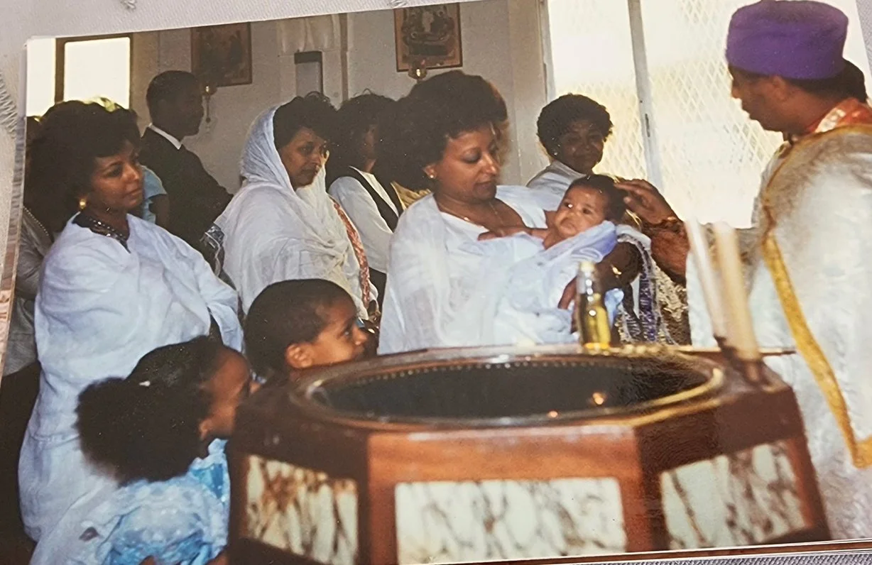 A woman holding a baby during a baptism ceremony inside a church, surrounded by women and children dressed in white, with a priest officiating.